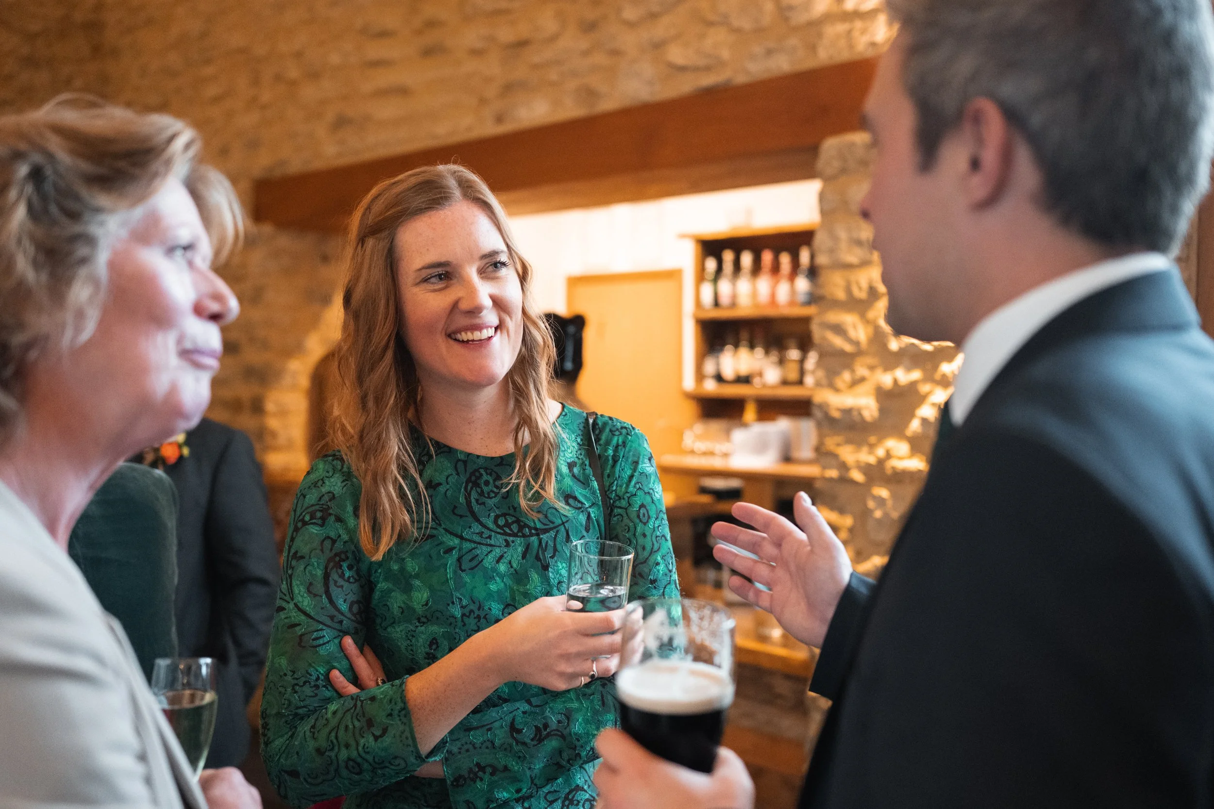 Group of people socializing indoors, woman in green dress smiling and holding a drink, man in suit and tie holding a dark beer, woman with blonde curly hair in beige blazer, in a cozy setting with brick walls and wooden accents.