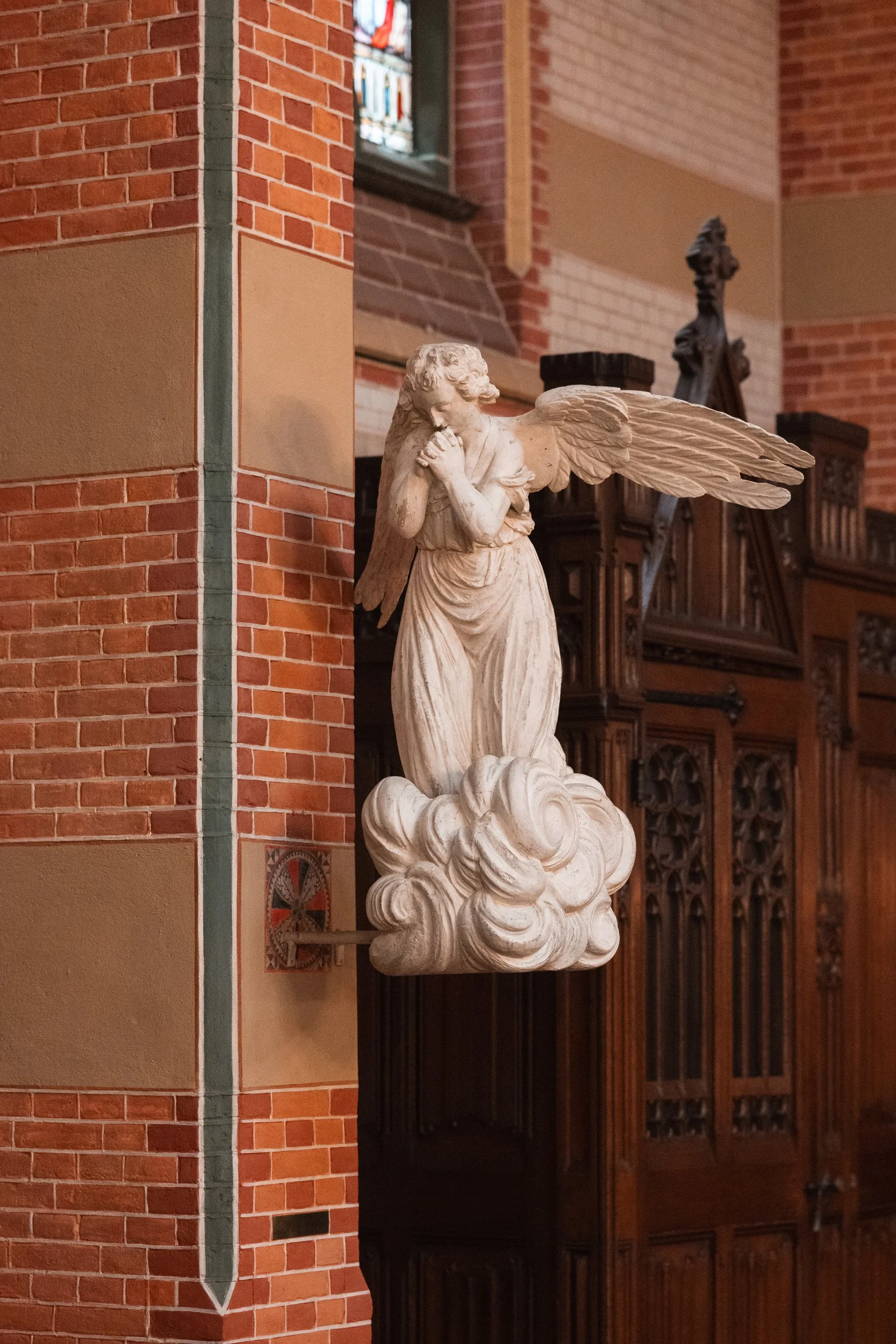 Statue of an angel with wings, standing on a cloud, inside a church with brick walls and stained glass window.