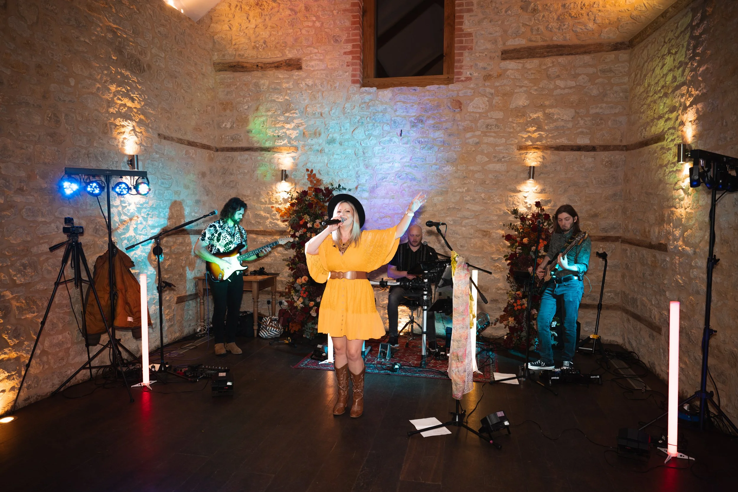 A woman singing into a microphone with a band playing behind her in a cozy venue with stone walls and colorful lighting.