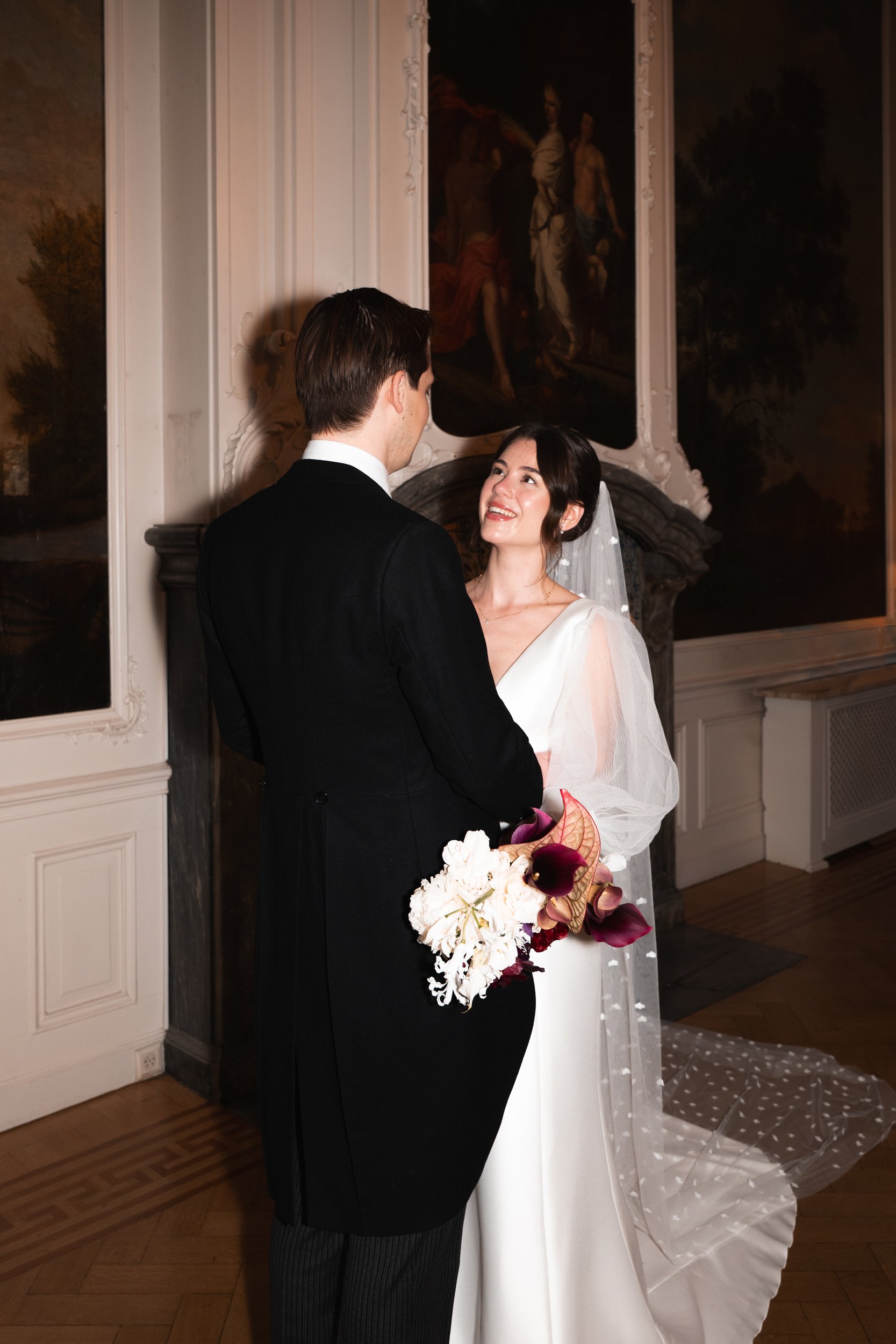 A bride and groom during their wedding ceremony in an elegant room, with the bride holding a bouquet of white and dark purple flowers, looking at each other happily.