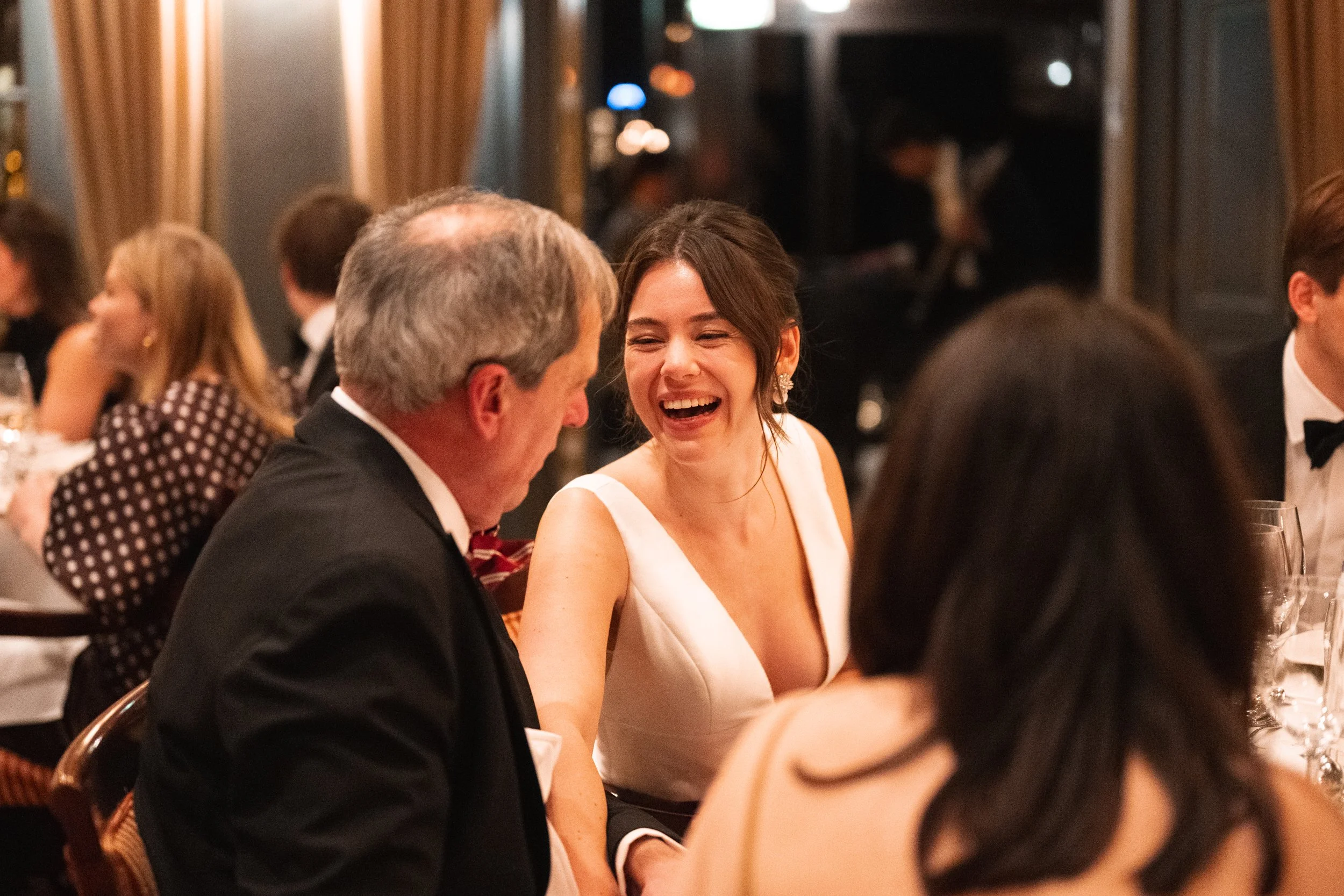 A woman laughing and smiling while talking to a man at a formal dinner event. Both are dressed in formal attire, with other guests in suits and dresses visible in the background.