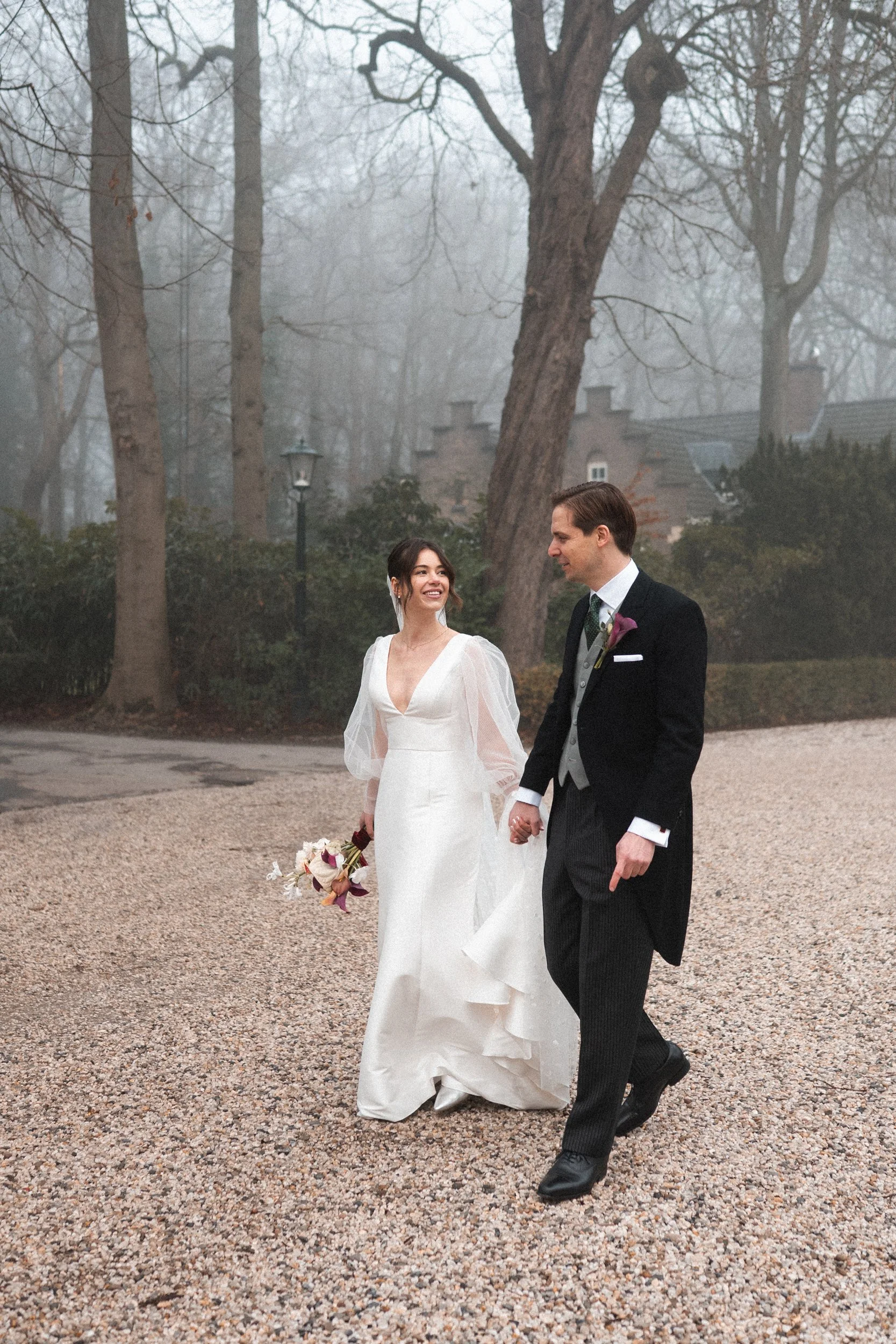 A bride and groom walking hand in hand outdoors in a misty, wooded area on their wedding day. The bride is holding a bouquet and wearing a white wedding gown, while the groom is dressed in a black tuxedo with a gray vest and a purple boutonniere.