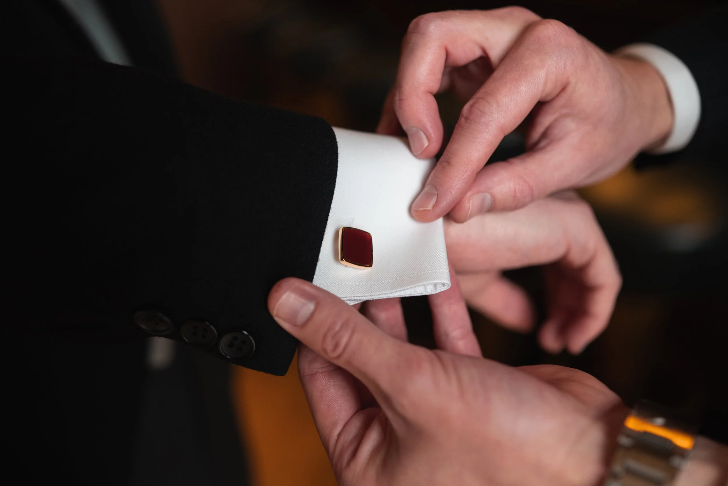 Person adjusting the cuff of another person's dress shirt with a red and gold cufflink, both wearing formal black suits.