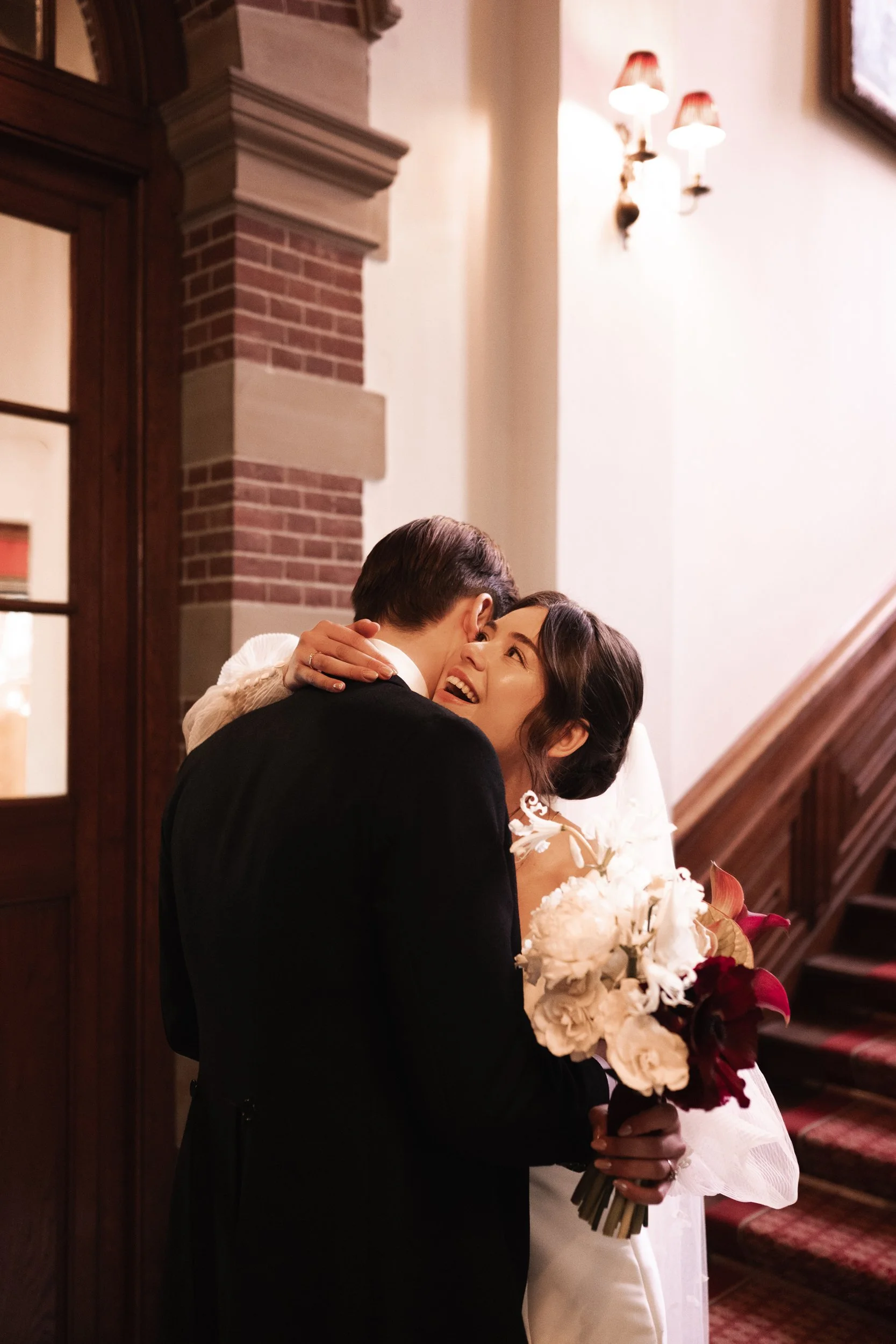 A joyful bride and groom hugging on their wedding day, with the bride holding a bouquet of white and red flowers, standing inside a warmly lit venue near stairs.