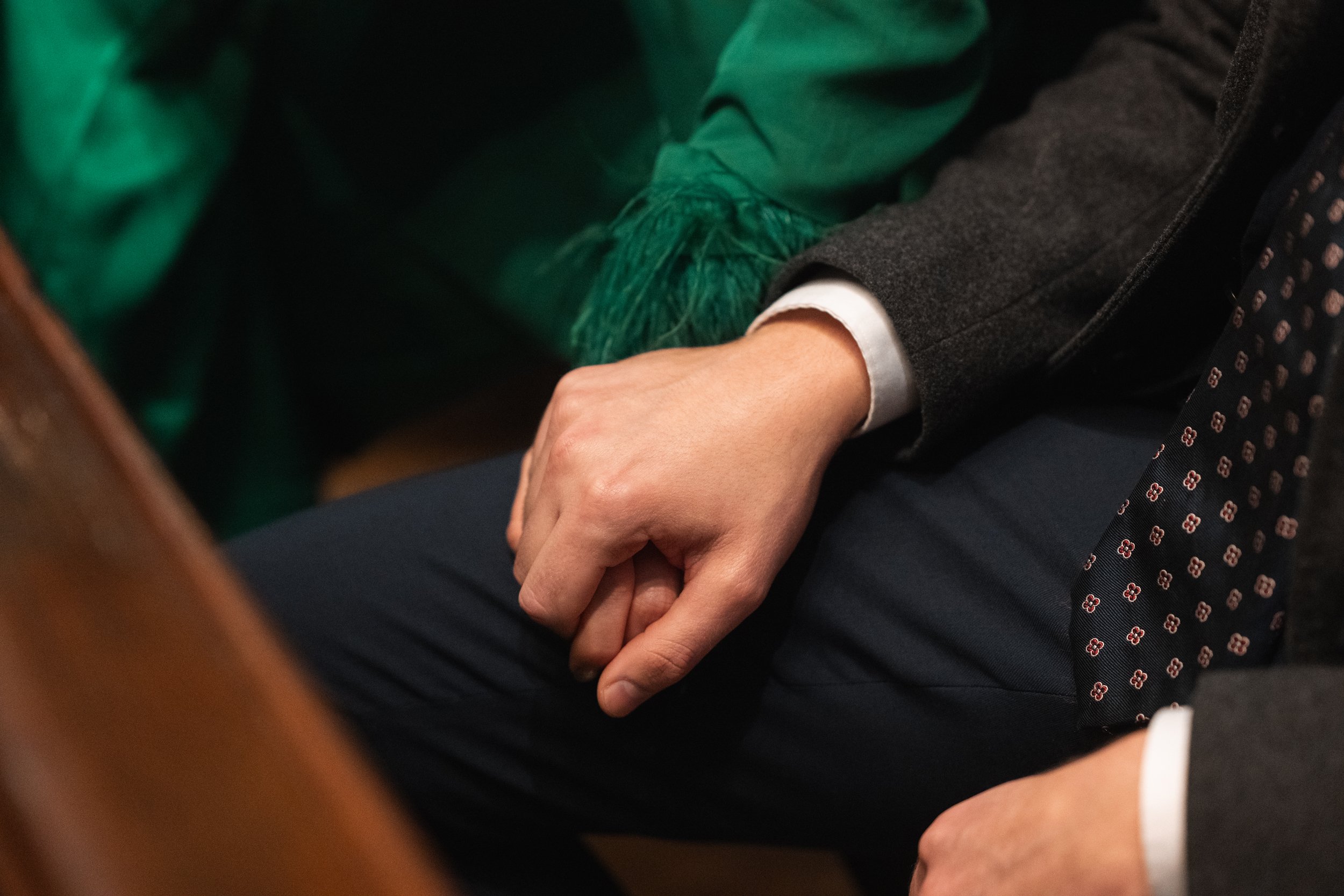 Close-up of a person wearing a dark gray suit with a white shirt, sitting with hands clasped on their lap. The person is seated on a wooden bench or pew, with a green fabric and another person partly visible wearing a dark suit and a dotted tie.