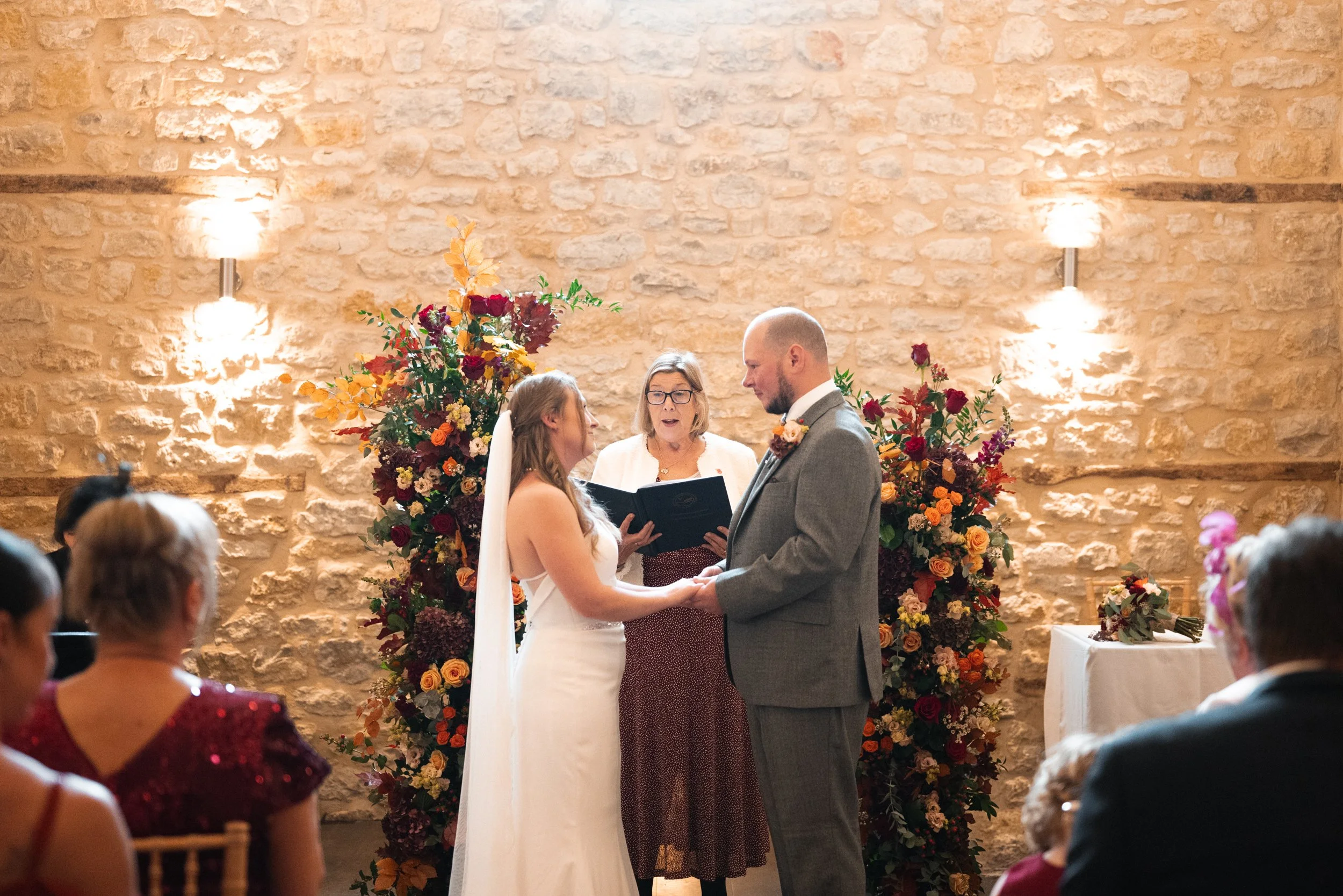 A wedding ceremony with a bride and groom holding hands, standing in front of an officiant with large floral arrangements behind them, inside a rustic stone-walled venue.