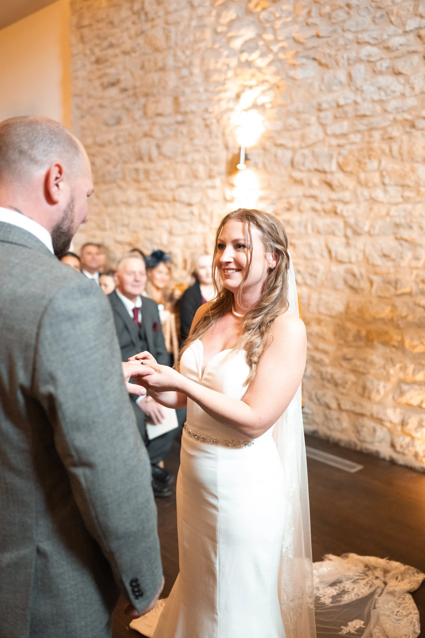 A bride and groom exchange rings during their wedding ceremony in a rustic venue with a stone wall and warm lighting. Guests are seated in the background, smiling and observing the moment.