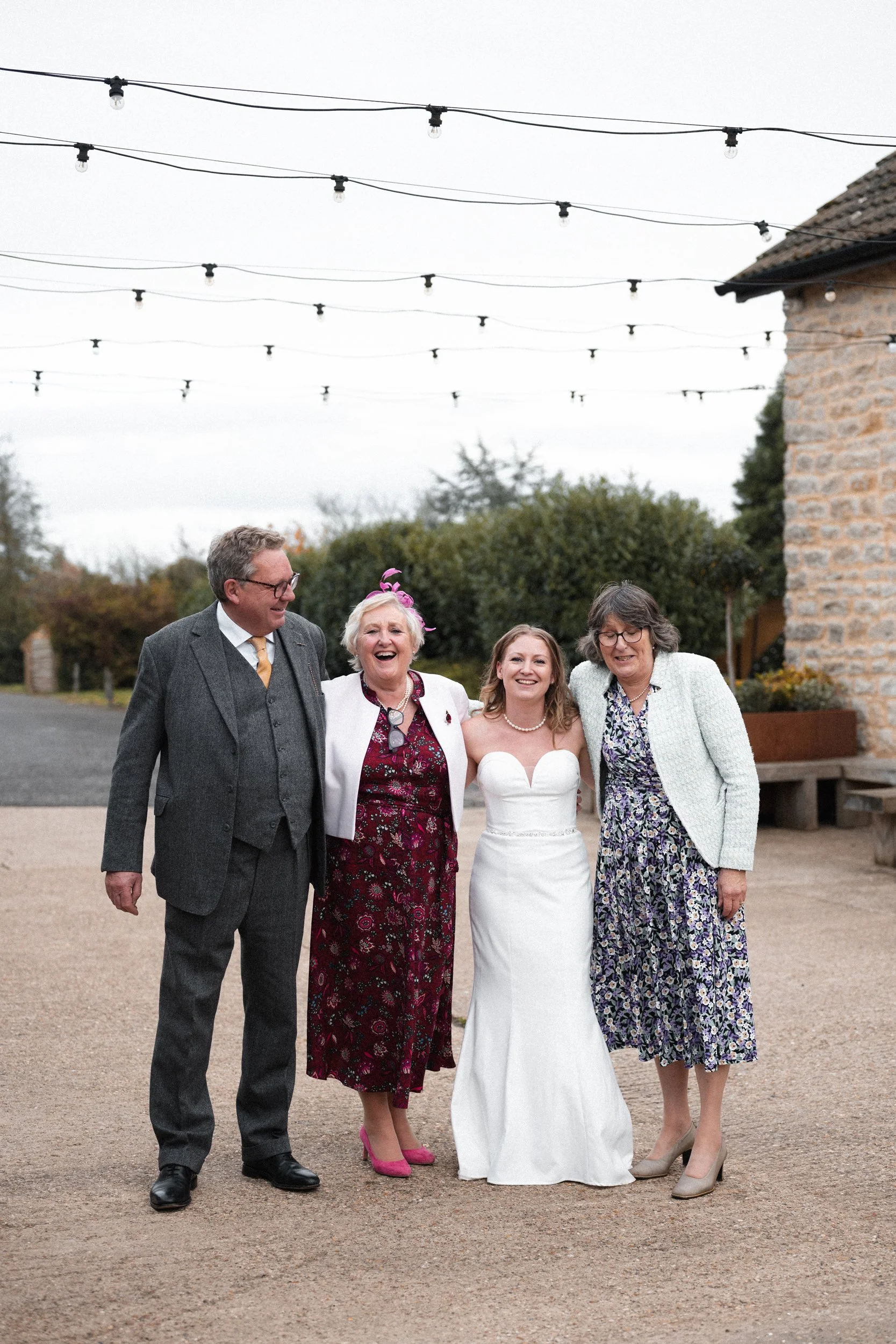 Four women and one man standing outdoors, hugging and smiling. One woman is in a white wedding dress, indicating a wedding celebration.