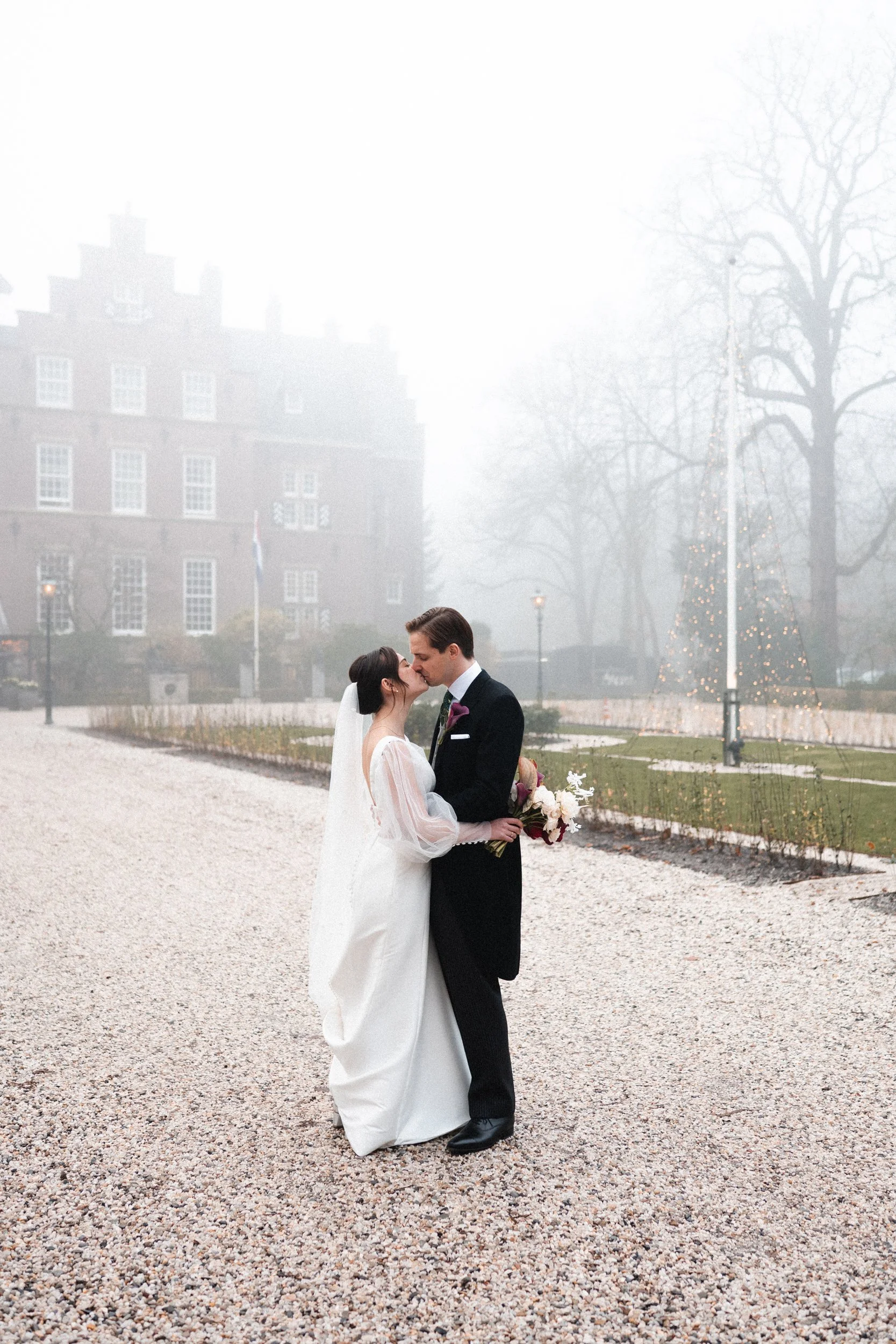 A bride and groom share a kiss outdoors on a gravel path, with a foggy background featuring a large building, leafless trees, and a decorated Christmas tree.