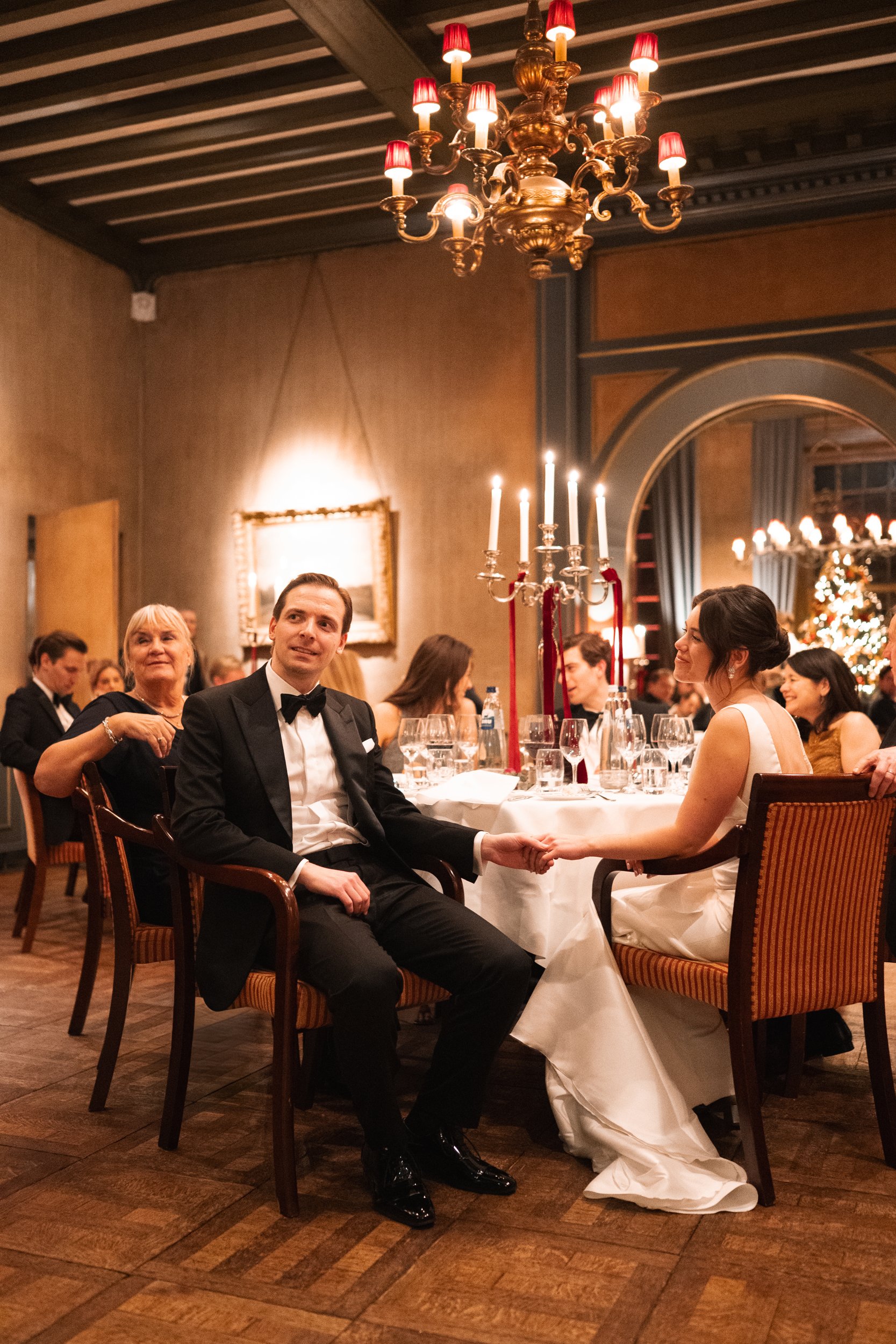 A wedding reception with a bride and groom holding hands at their table, surrounded by guests in formal attire, decorated with chandeliers, candelabras, and a Christmas tree in the background.