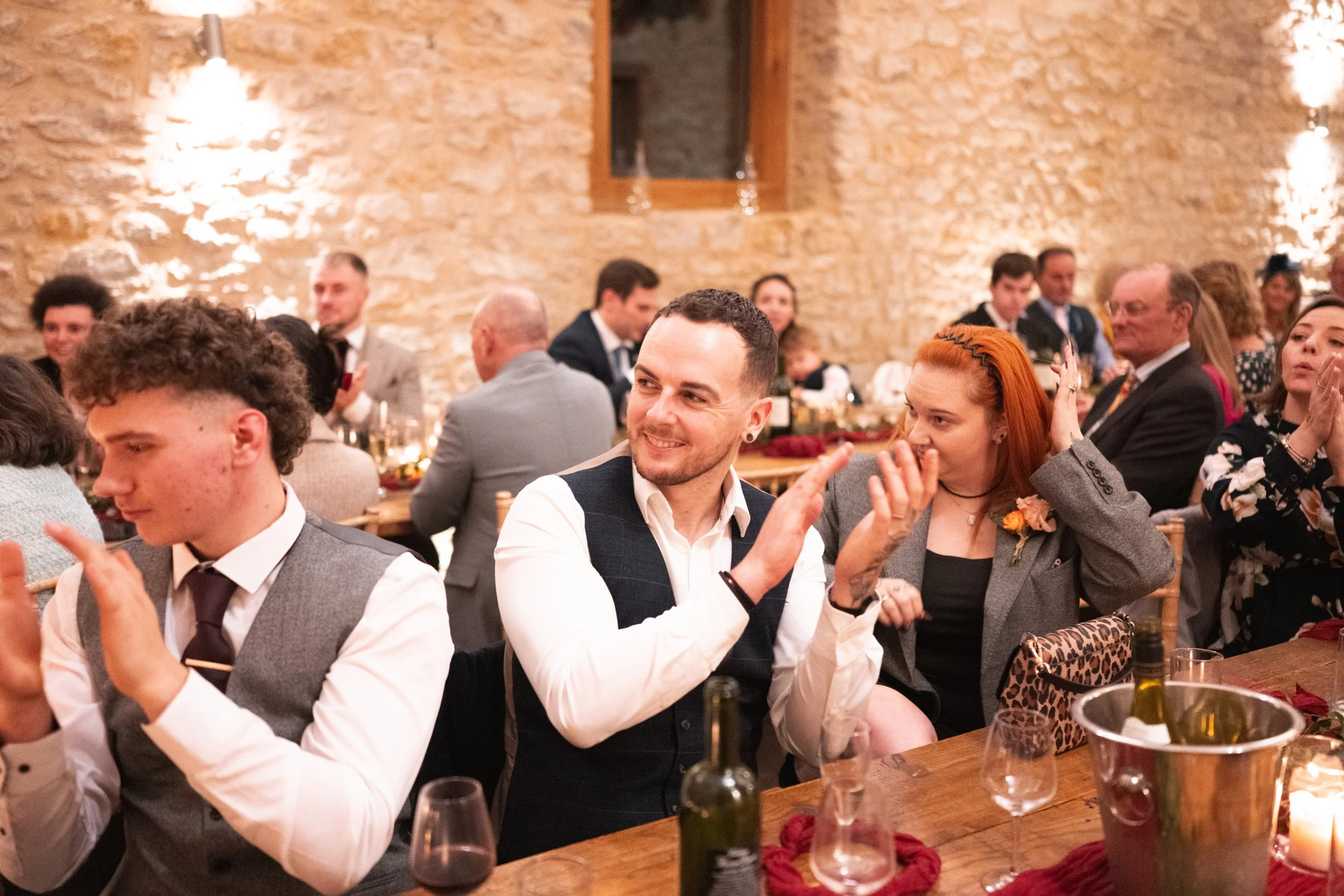 People at a formal event clapping and socializing in a warmly lit brick-walled room with wooden tables and chairs.