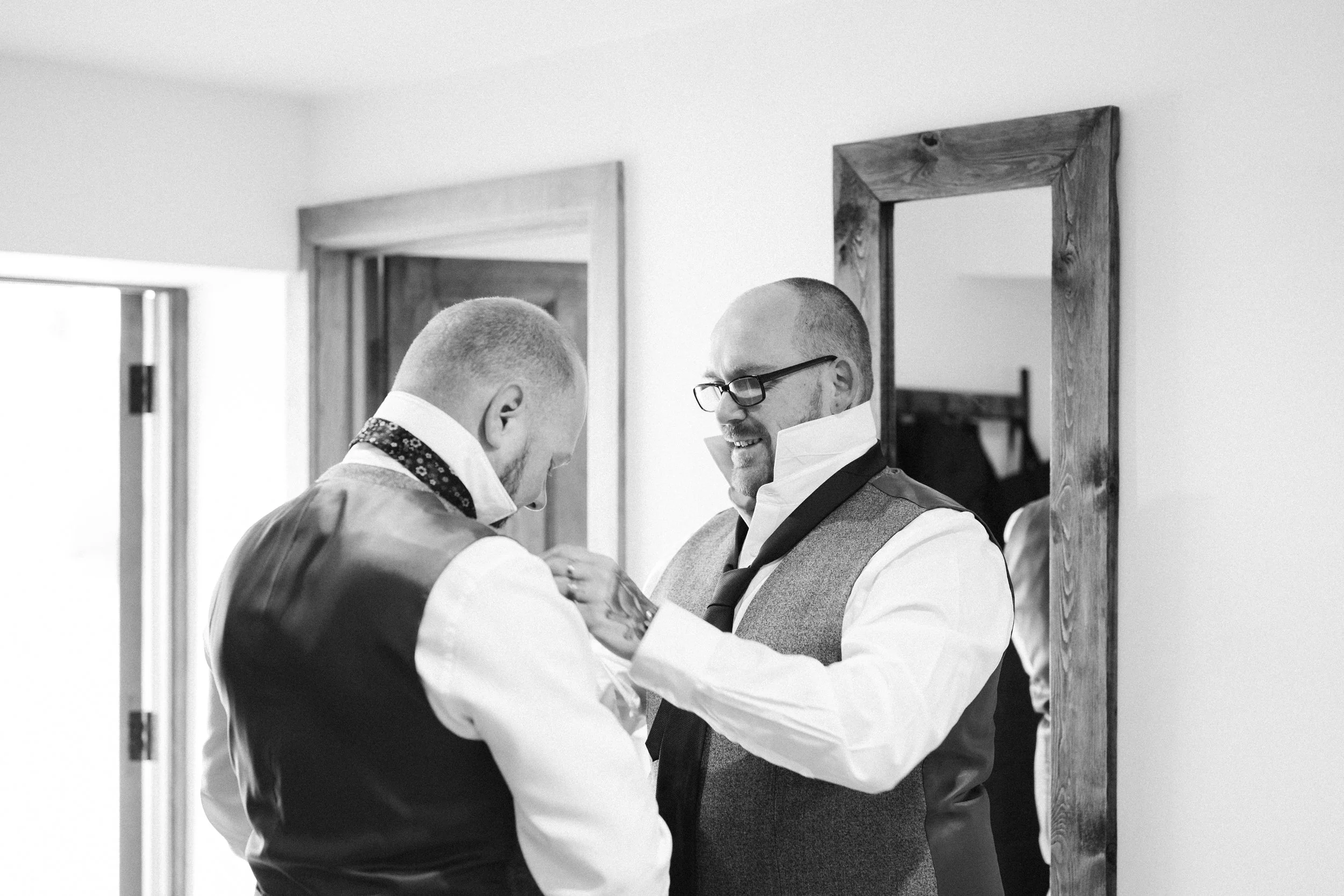 Two men getting ready, one adjusting the other's collar in a mirror, both dressed in formal attire, in a room with wooden frames and a mirror.