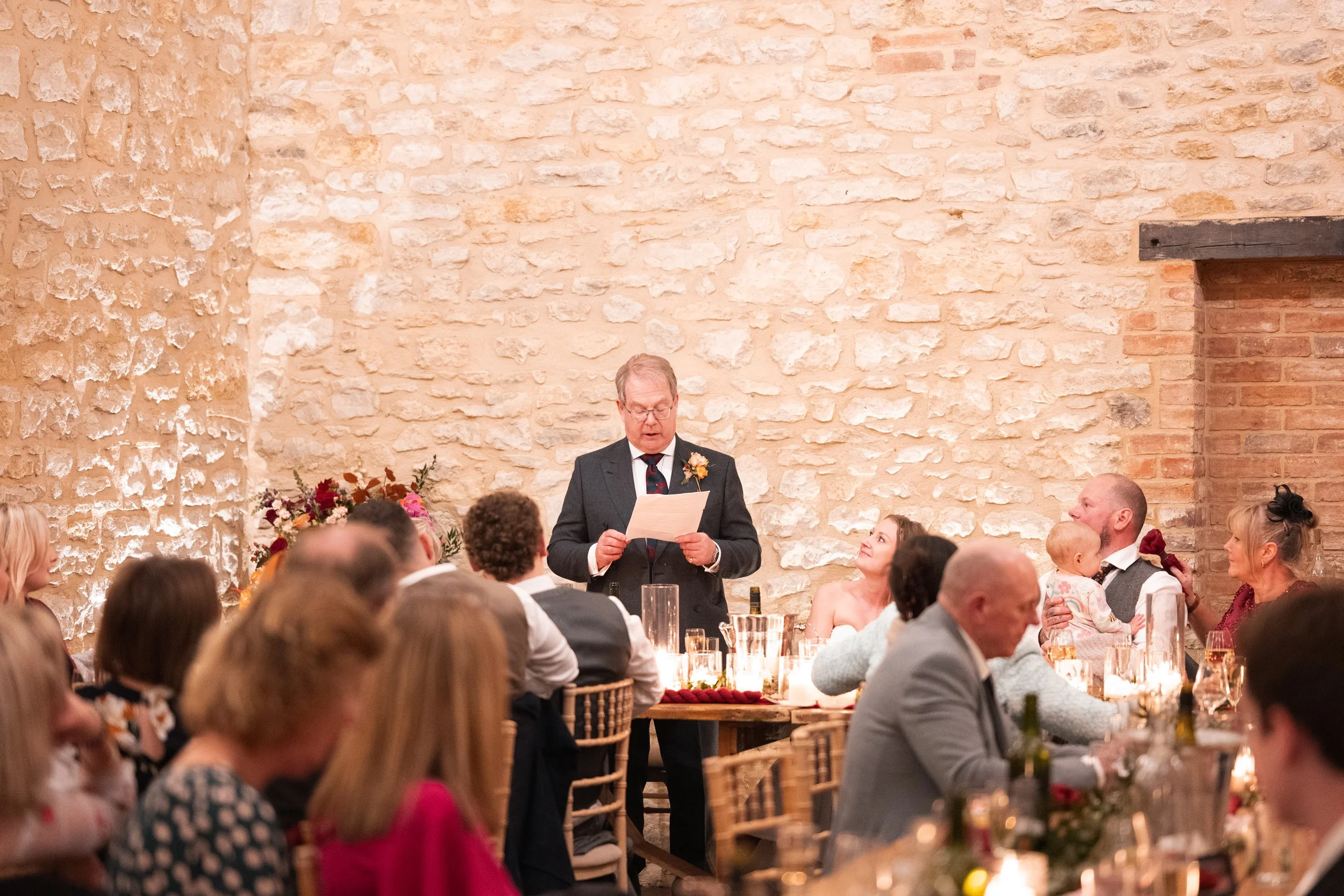 A man giving a speech at a wedding reception, surrounded by seated guests in a rustic venue with exposed brick walls, candlelit tables, and floral decorations.