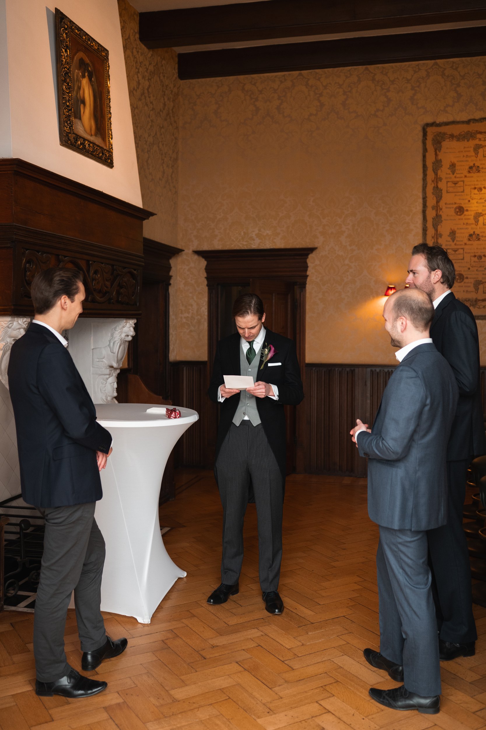 A group of four men dressed in suits standing in a room, with one man in the center reading a card. The room has wood-paneled walls, a fireplace with ornamental carving, a painting of a woman on the wall, and a tall round table with a couple of dice 