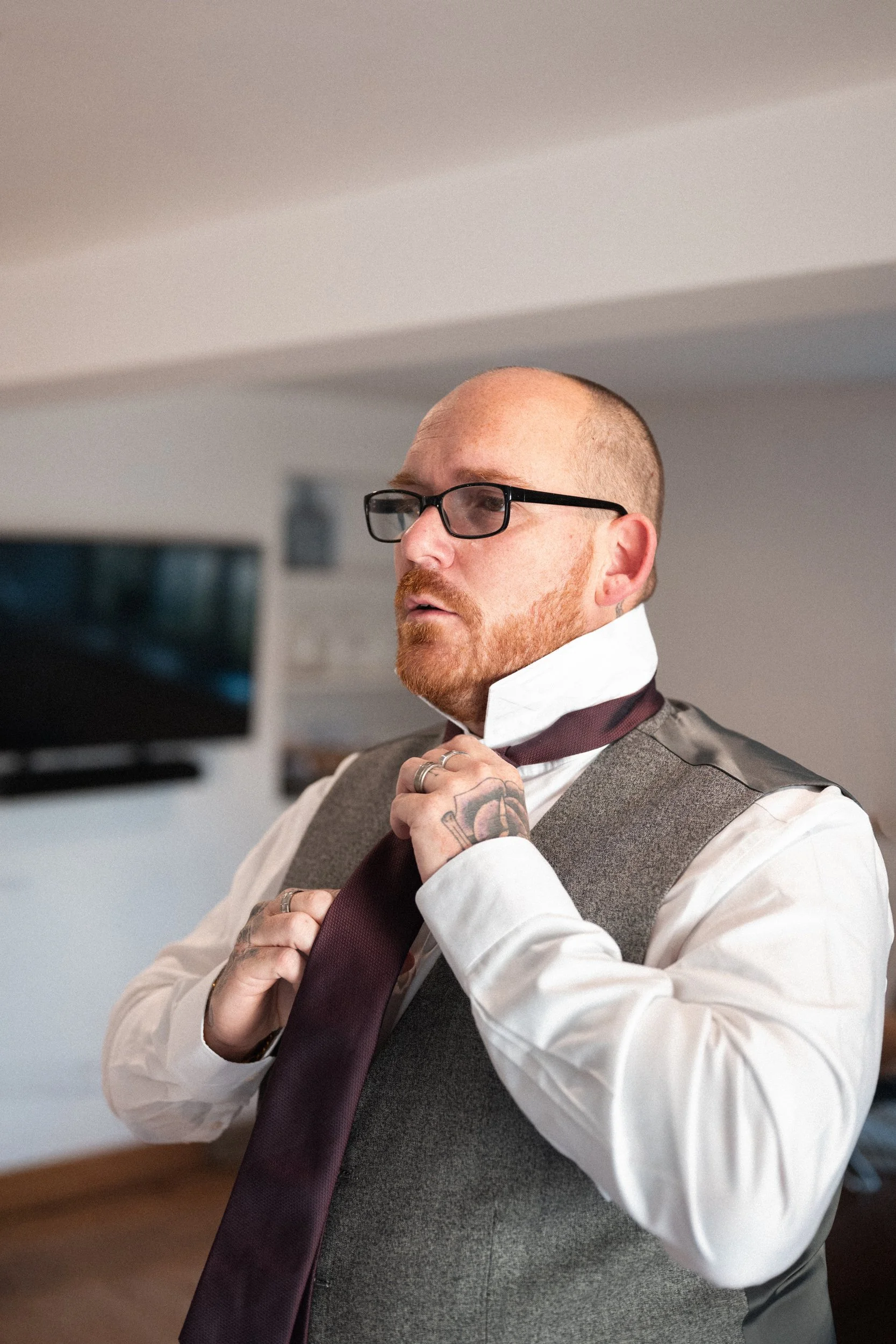 Man with glasses adjusting a tie, wearing a white shirt and gray vest, in a room with a television in the background.