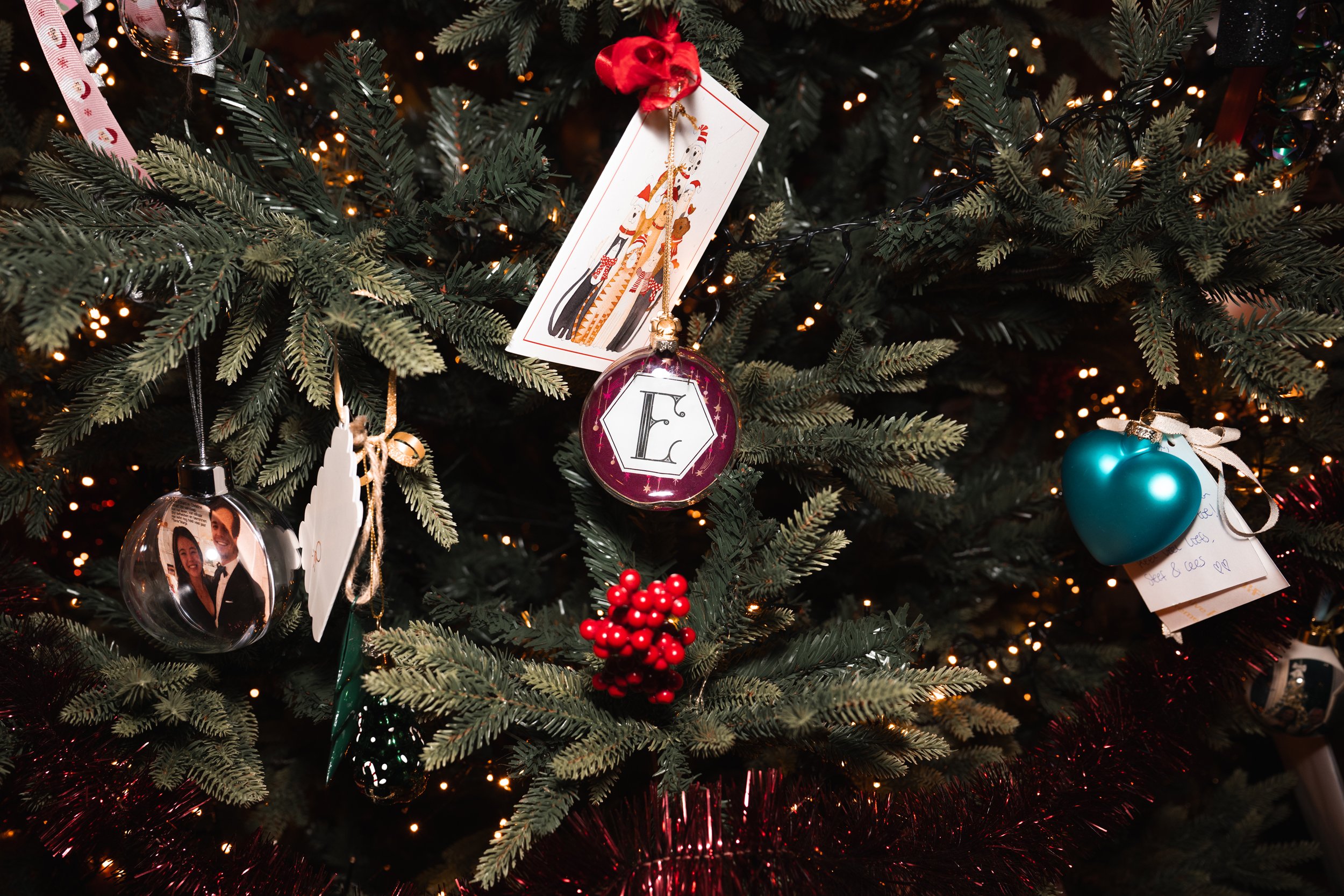Close-up of a decorated Christmas tree with various ornaments, including a photo ornament, an monogrammed ornament, a heart-shaped ornament, and a berry cluster, with warm lights and festive decorations.