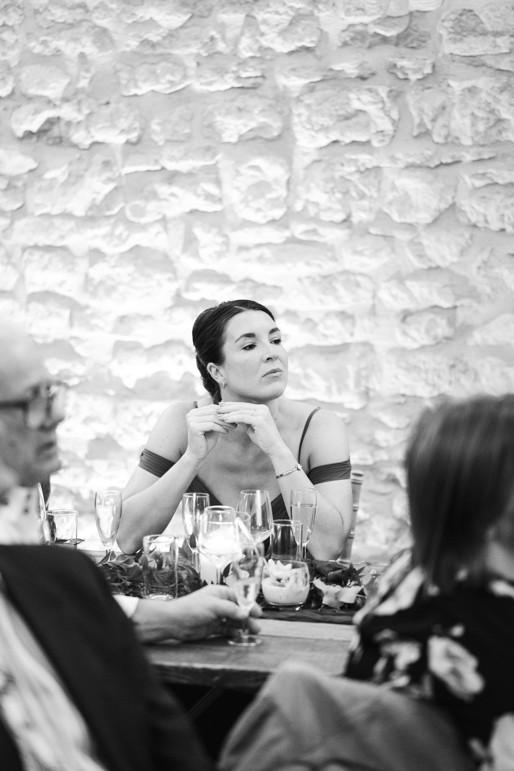 A woman with dark hair styled back sits at a table, looking thoughtfully to the side. She is surrounded by glasses and candles, with a textured stone wall in the background.