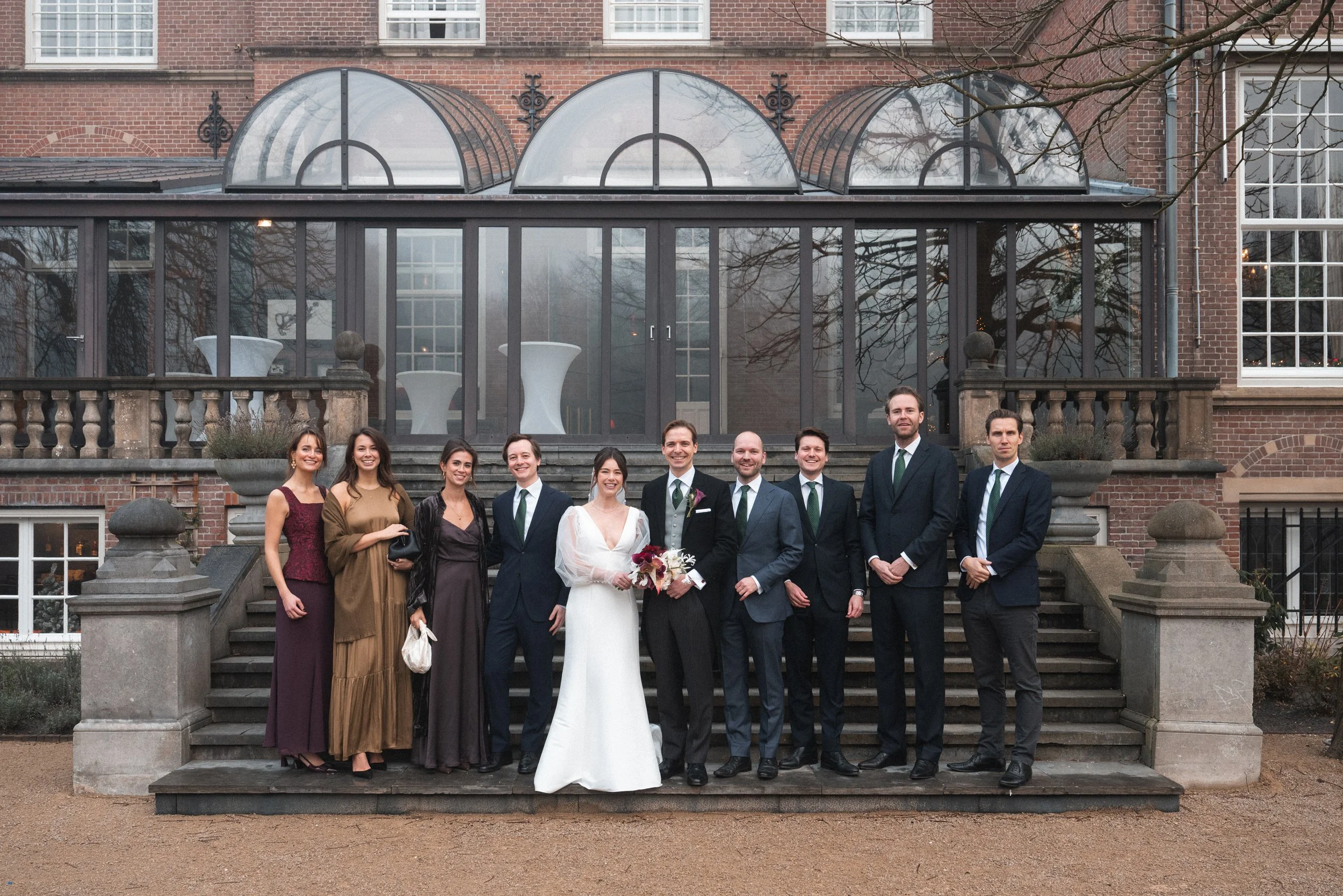 A wedding party standing on steps outside a brick building. The bride is in a white gown holding a bouquet, and the groom is in a suit with a gray vest and black tie. There are six men in suits and four women in dresses. The background features a gla