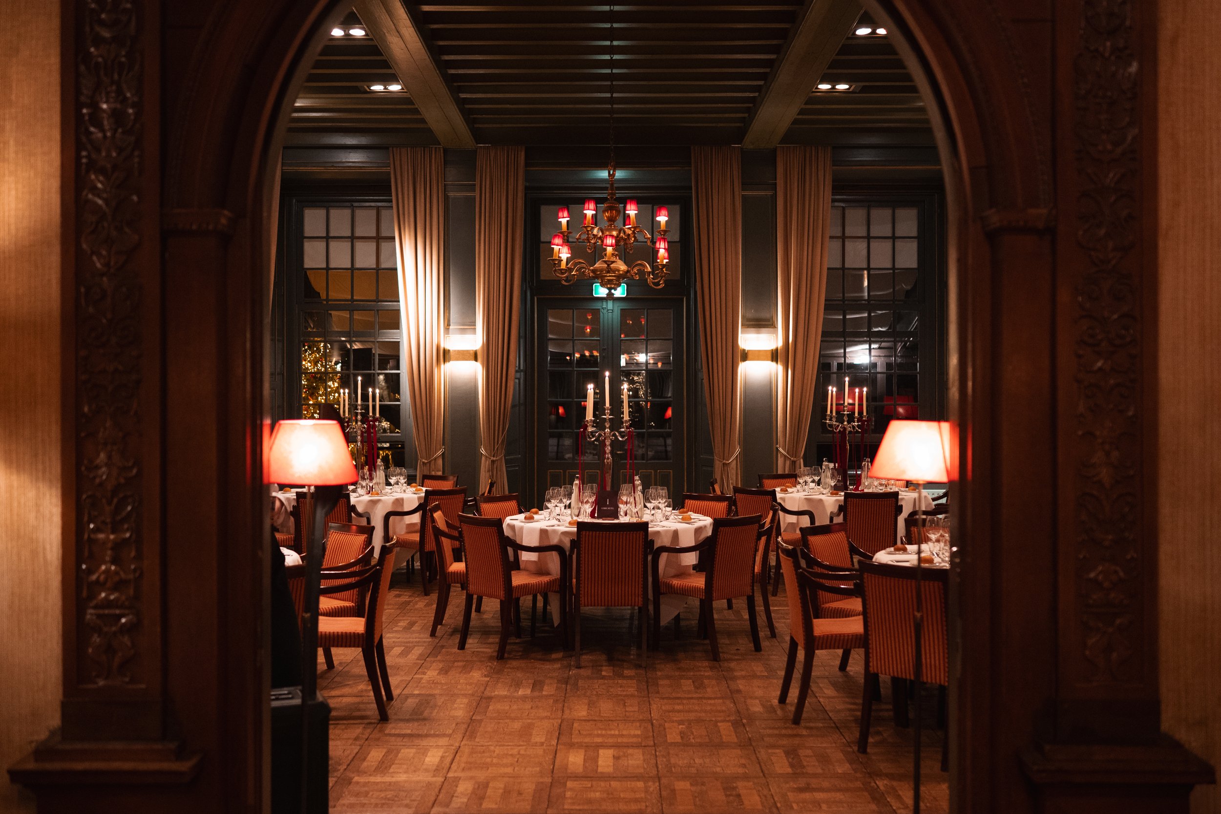 Elegant dining room with round tables covered in white tablecloths, surrounded by wooden chairs with striped upholstery. The room has warm lighting from table lamps and chandeliers, large windows with draped curtains, and candelabras on the tables, c