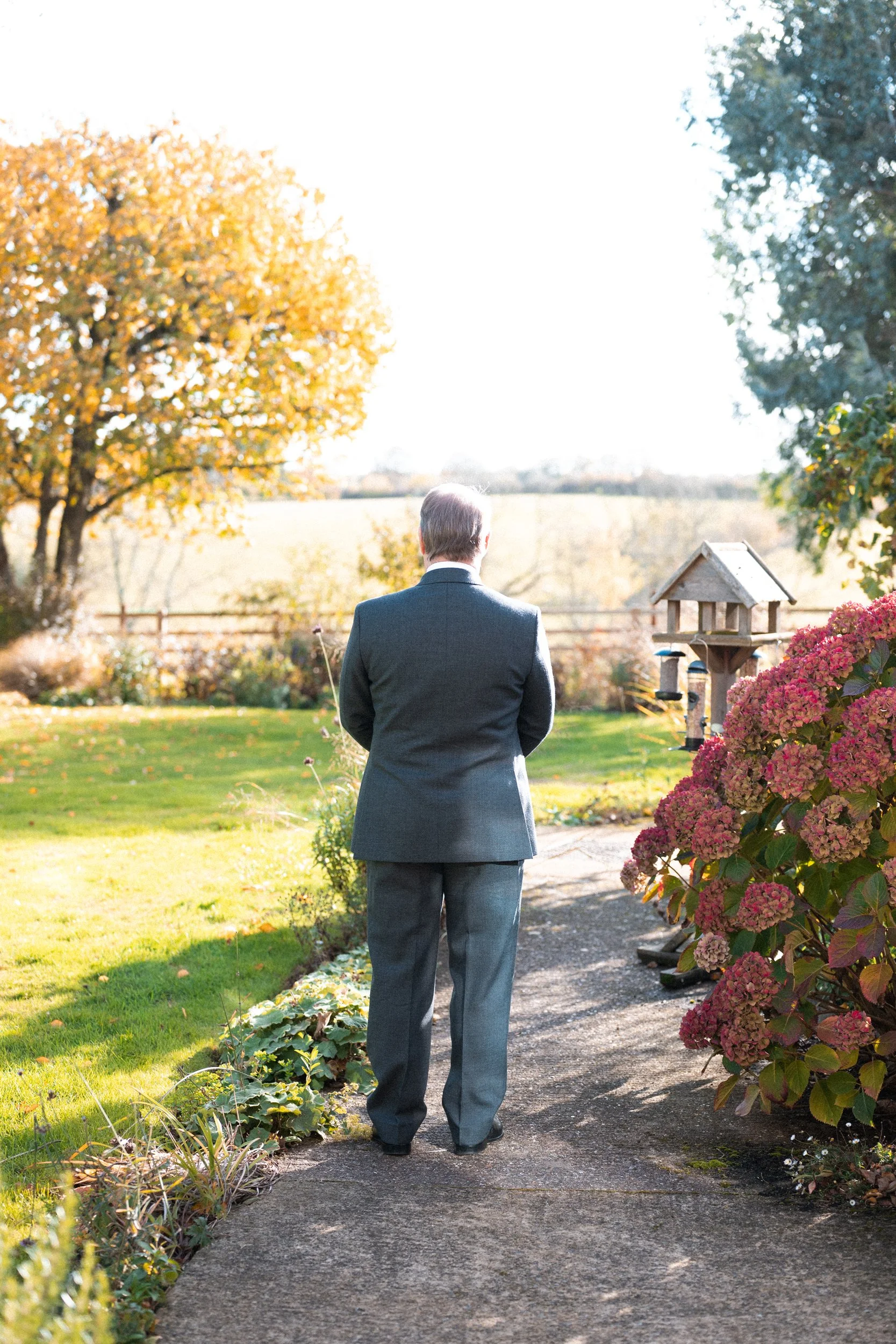 A man in a gray suit standing on a garden path, facing away, with a birdhouse and pink hydrangeas nearby and trees in the background.