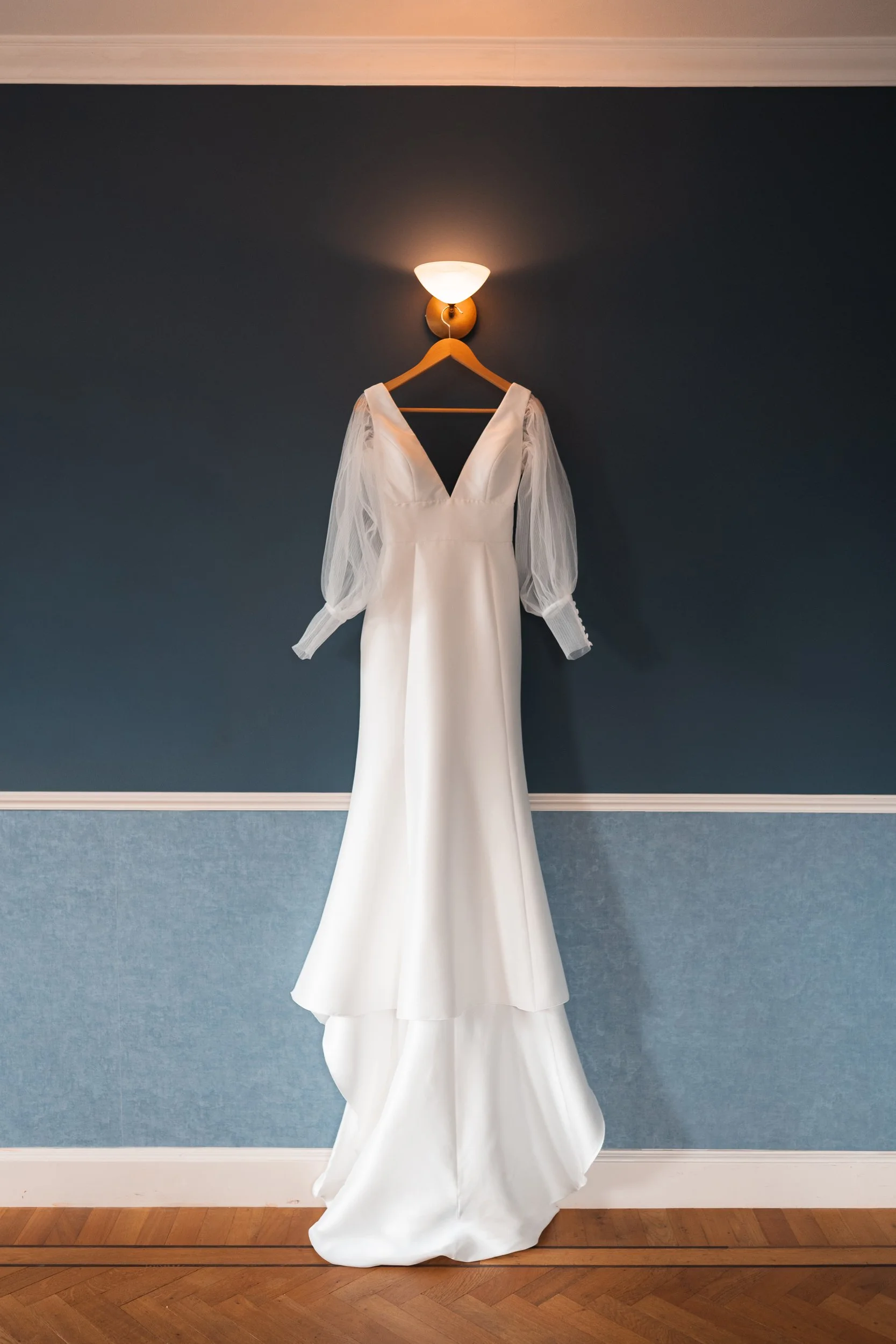 A wedding dress with puffed sheer long sleeves hanging on a wooden hanger, illuminated by a wall-mounted light fixture on a dark blue wall.