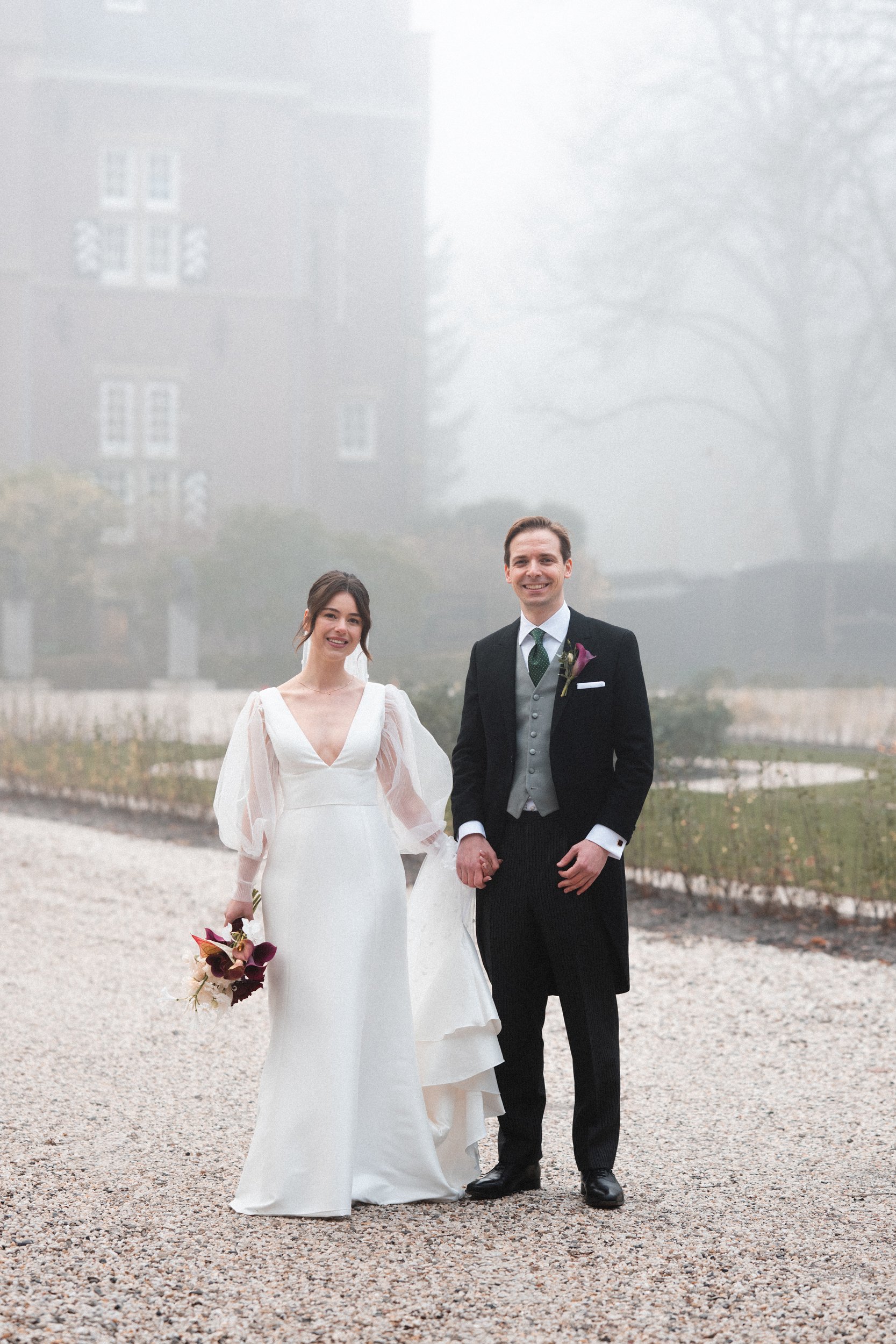 A newlywed couple stands hand in hand outdoors on a gravel path on a foggy day, with a large building and trees in the background.