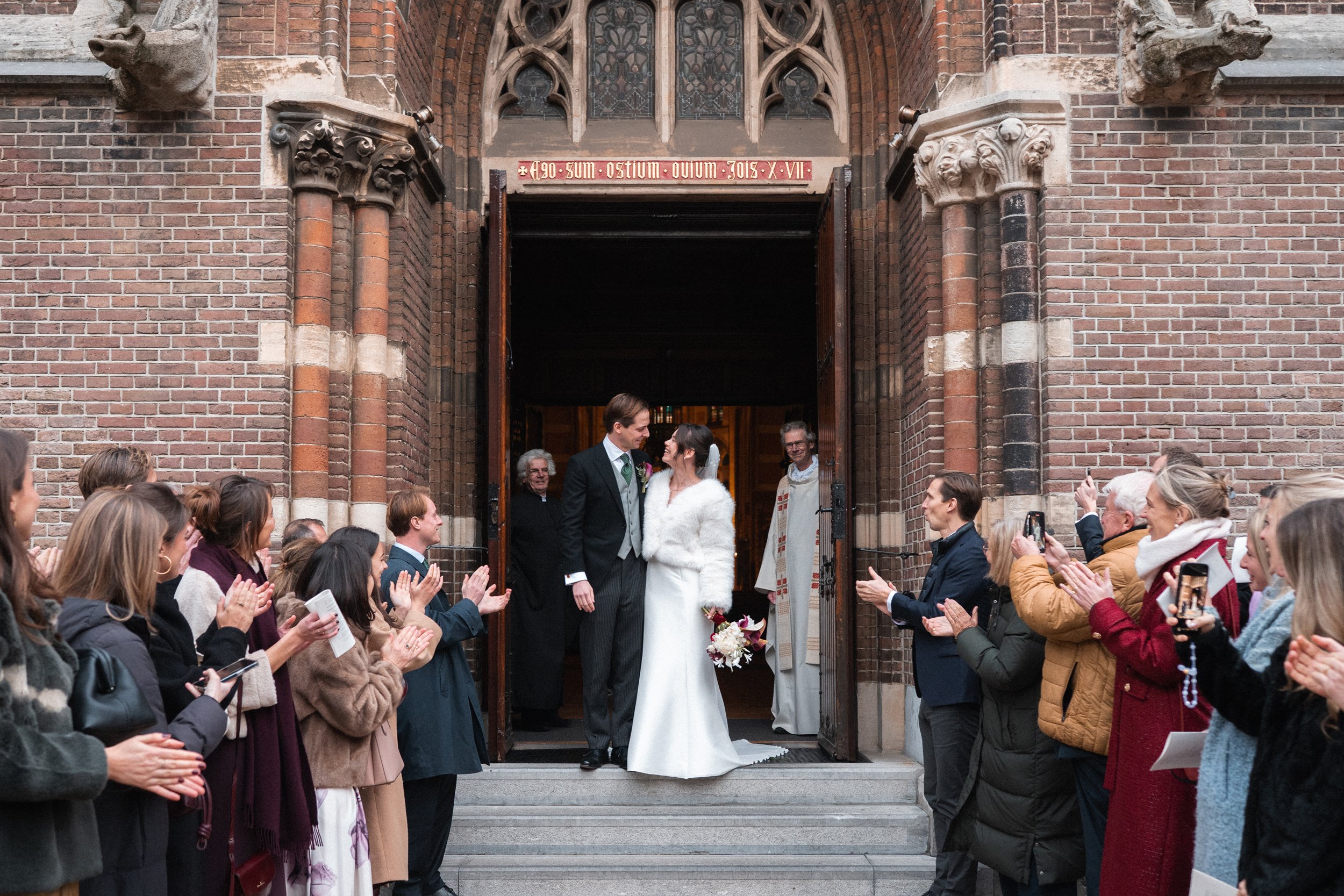 A wedding couple standing at the entrance of a church, looking at each other, surrounded by guests applauding and taking photos.