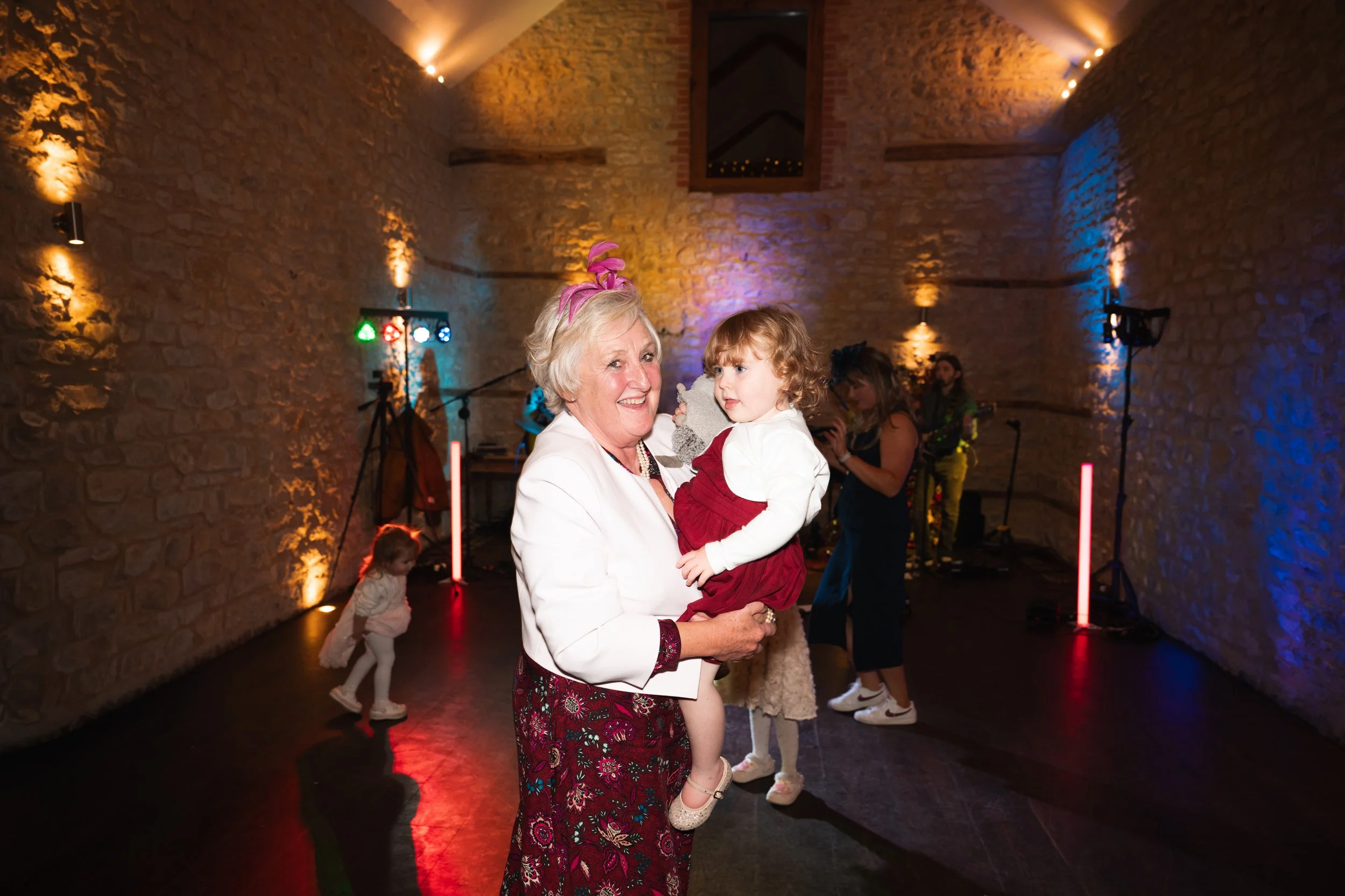 An elderly woman holding a young girl at a party or celebration in a dimly lit venue with colorful lighting, other children in the background, and a musical band performing.