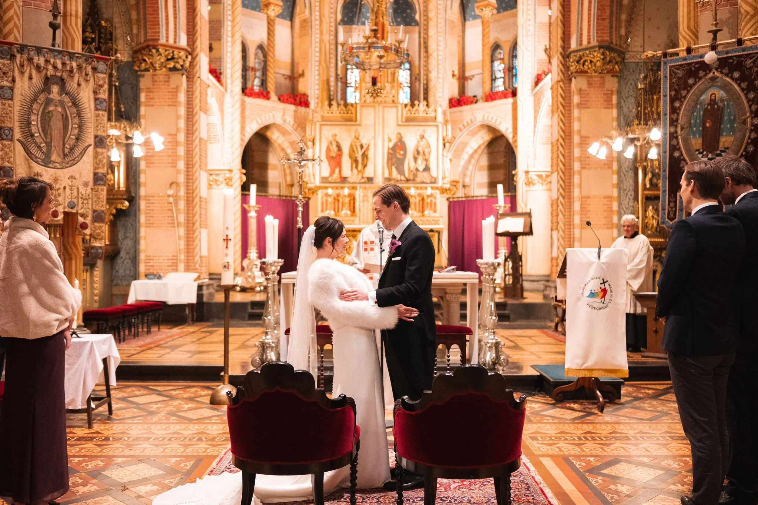 A wedding ceremony in a church with the bride and groom holding hands and facing each other, surrounded by guests and officiant, with ornate decorations and religious icons in the background.