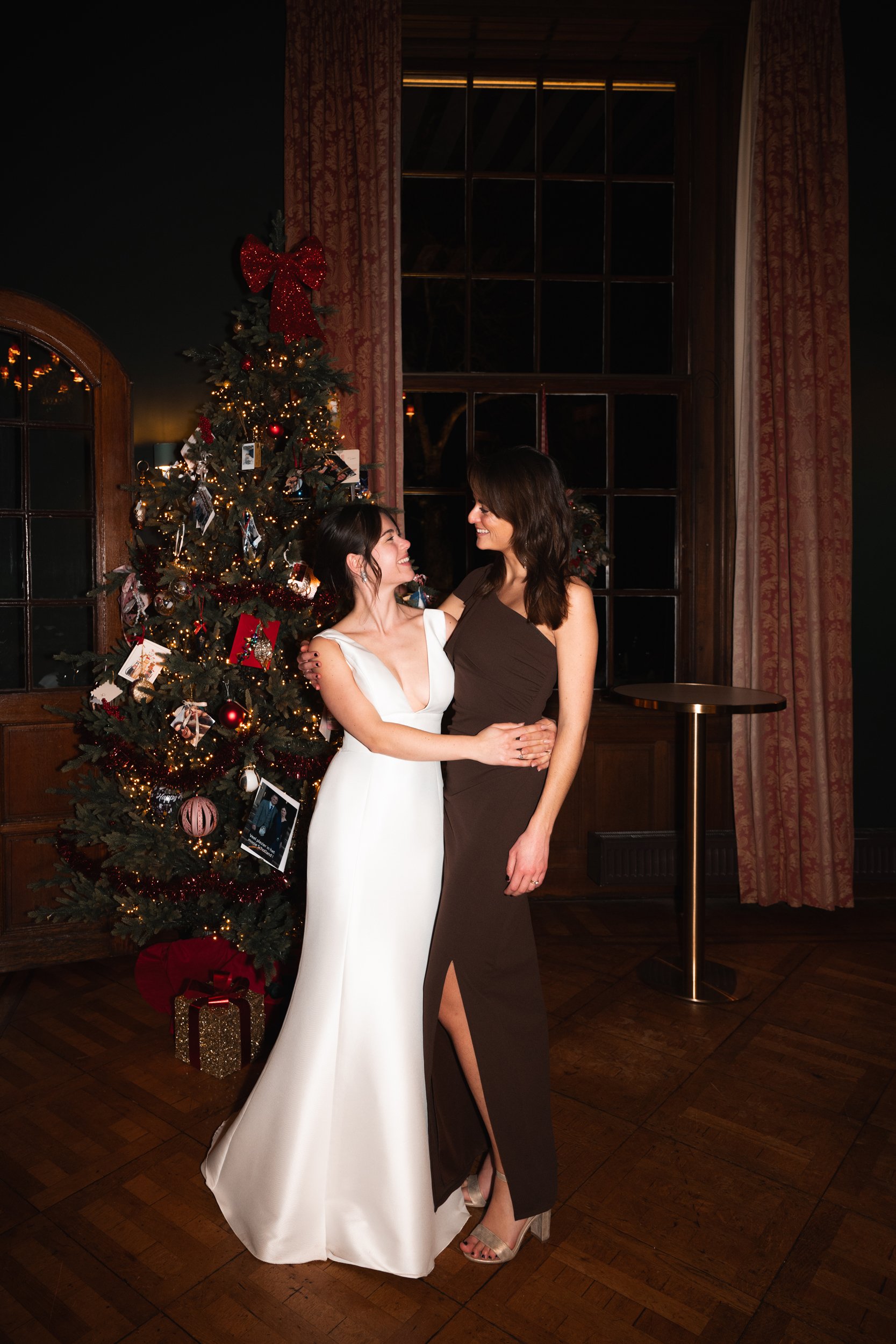 Two women in evening dresses embracing and smiling at each other in front of a decorated Christmas tree indoors.