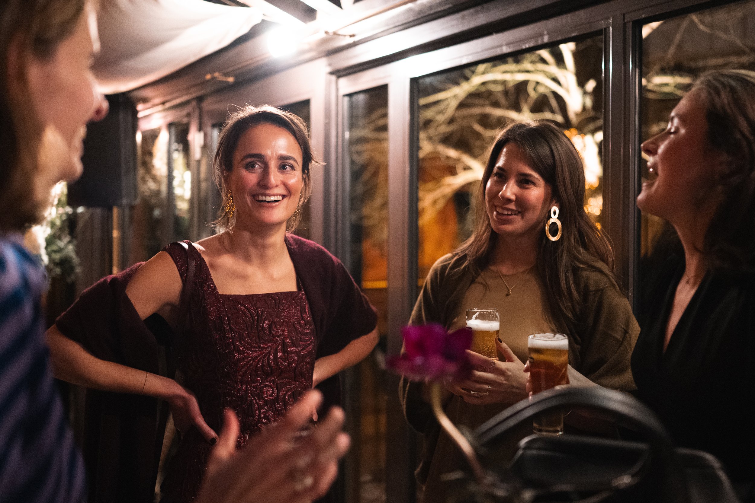 Four women socializing at a nighttime outdoor gathering, holding drinks and conversing inside near a large window with a view of trees outside.
