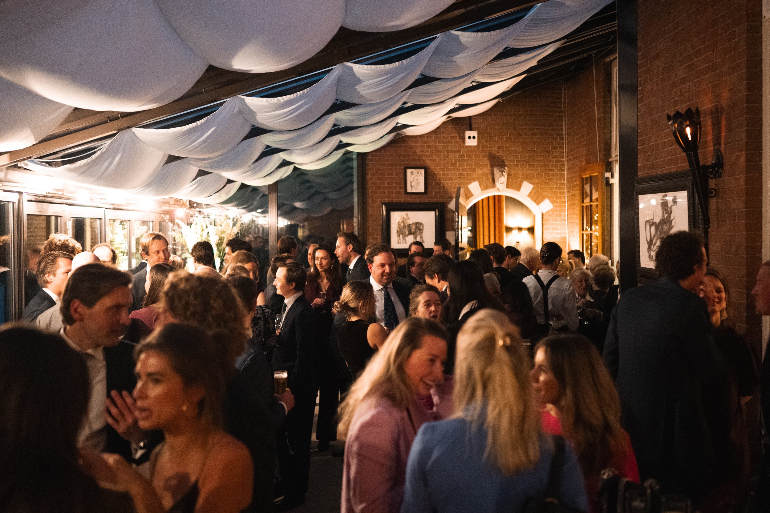 People socializing at an indoor party or event, dressed in formal attire, with decorated ceiling and warm lighting.