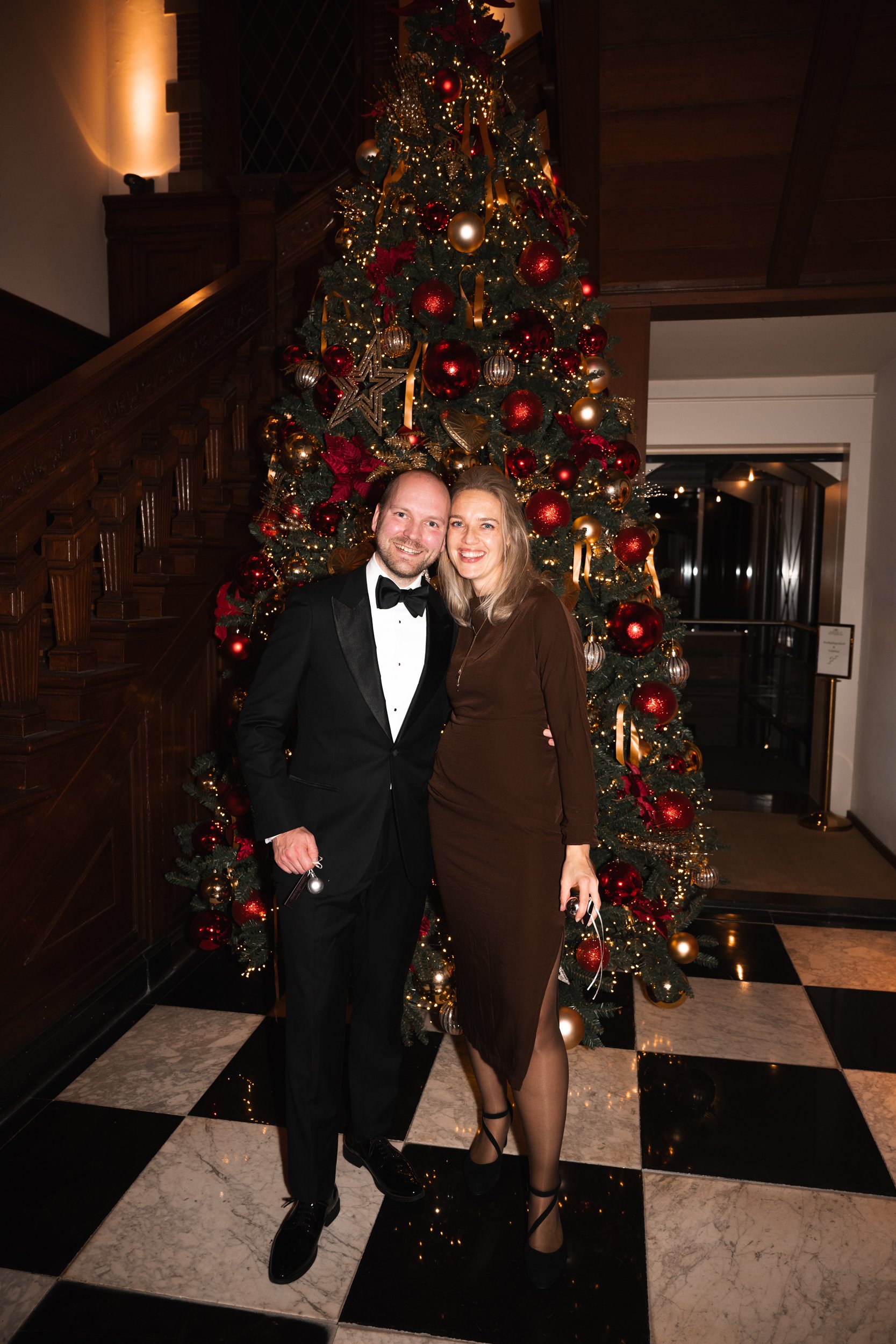 A man in a tuxedo and woman in a brown dress standing together in front of a decorated Christmas tree in an elegant setting.