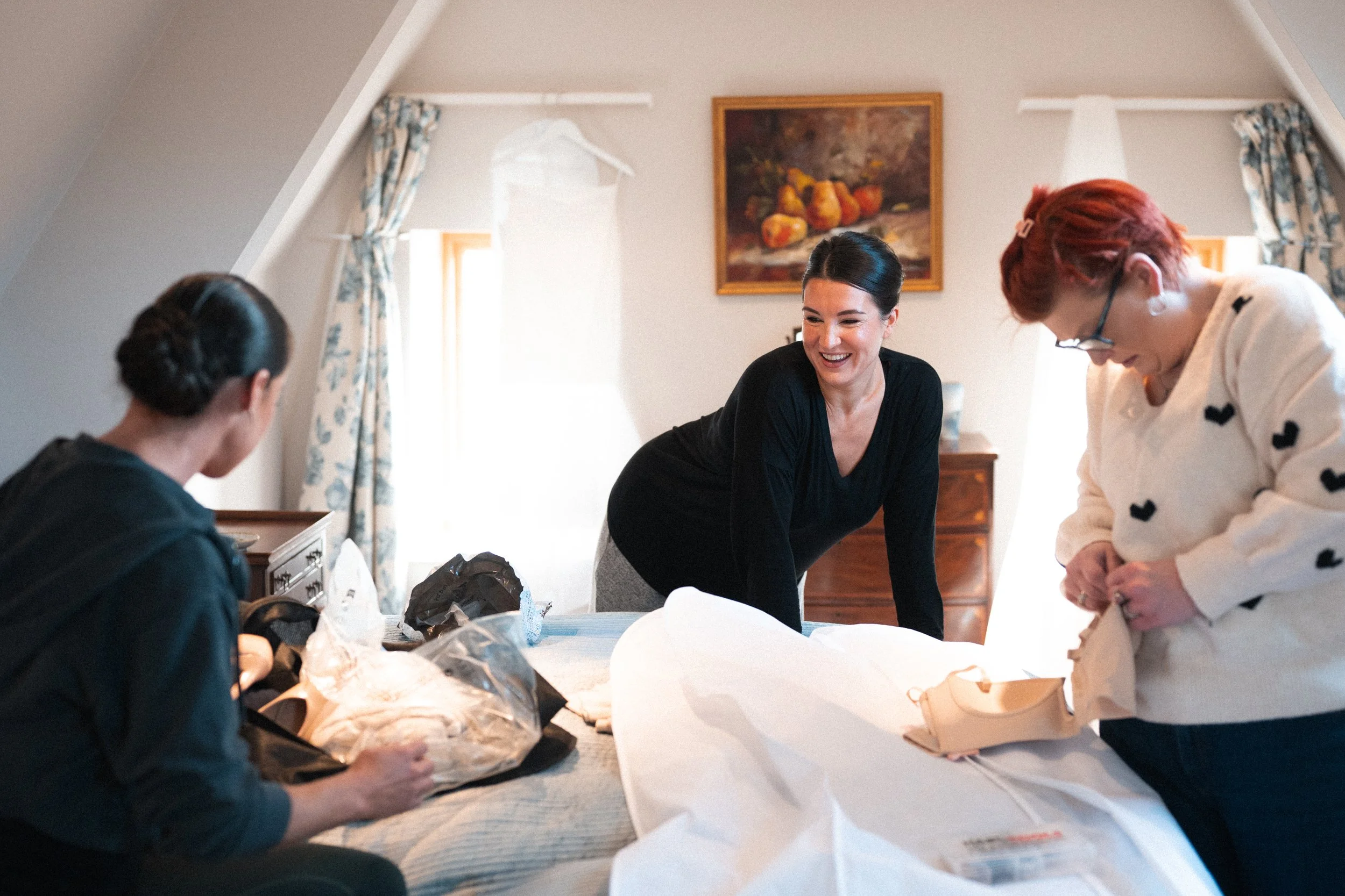Three women are sorting through clothing and shoes on a bed in a bright, cozy bedroom with sunlight streaming in through the windows.