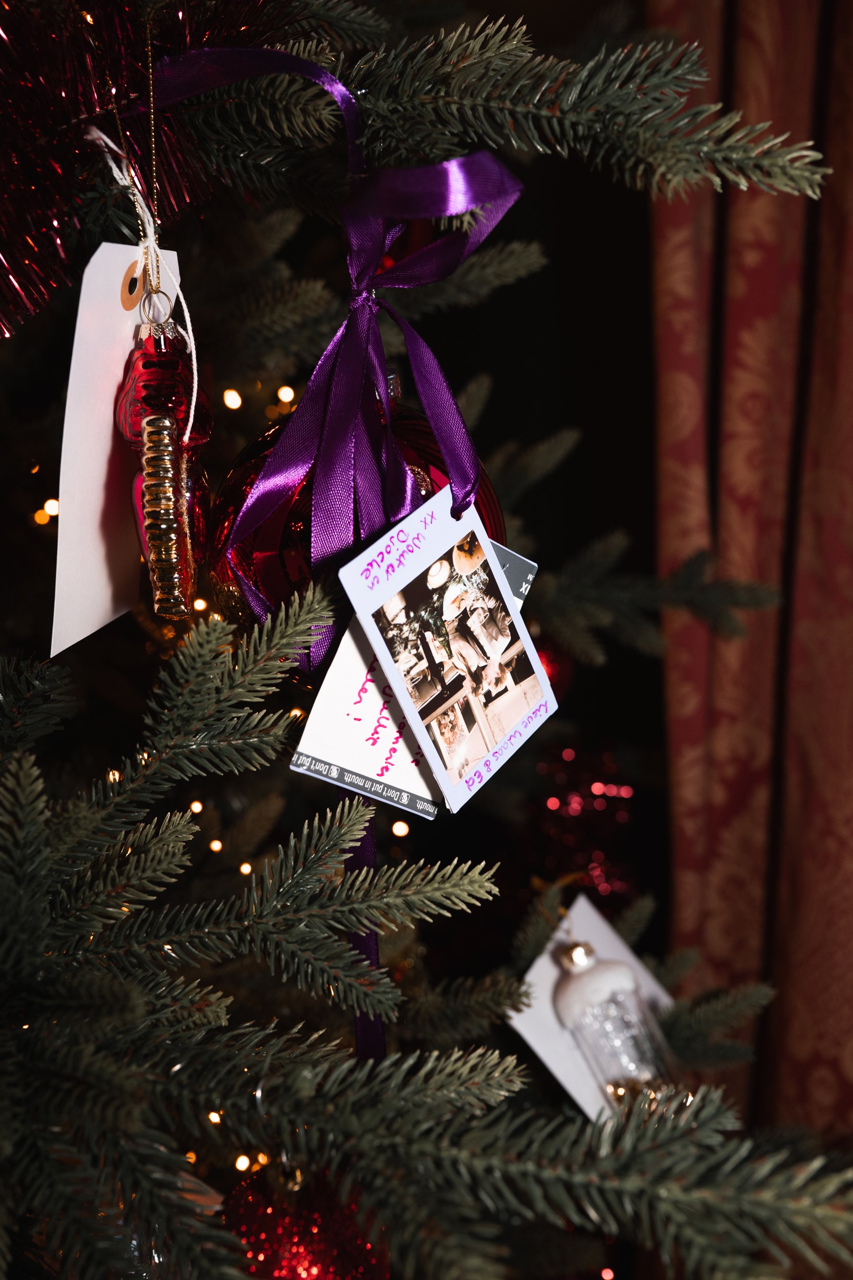 Close-up of a decorated Christmas tree with ornaments, ribbons, and small postcards hanging from the branches, with a dark background and a curtain to the right.