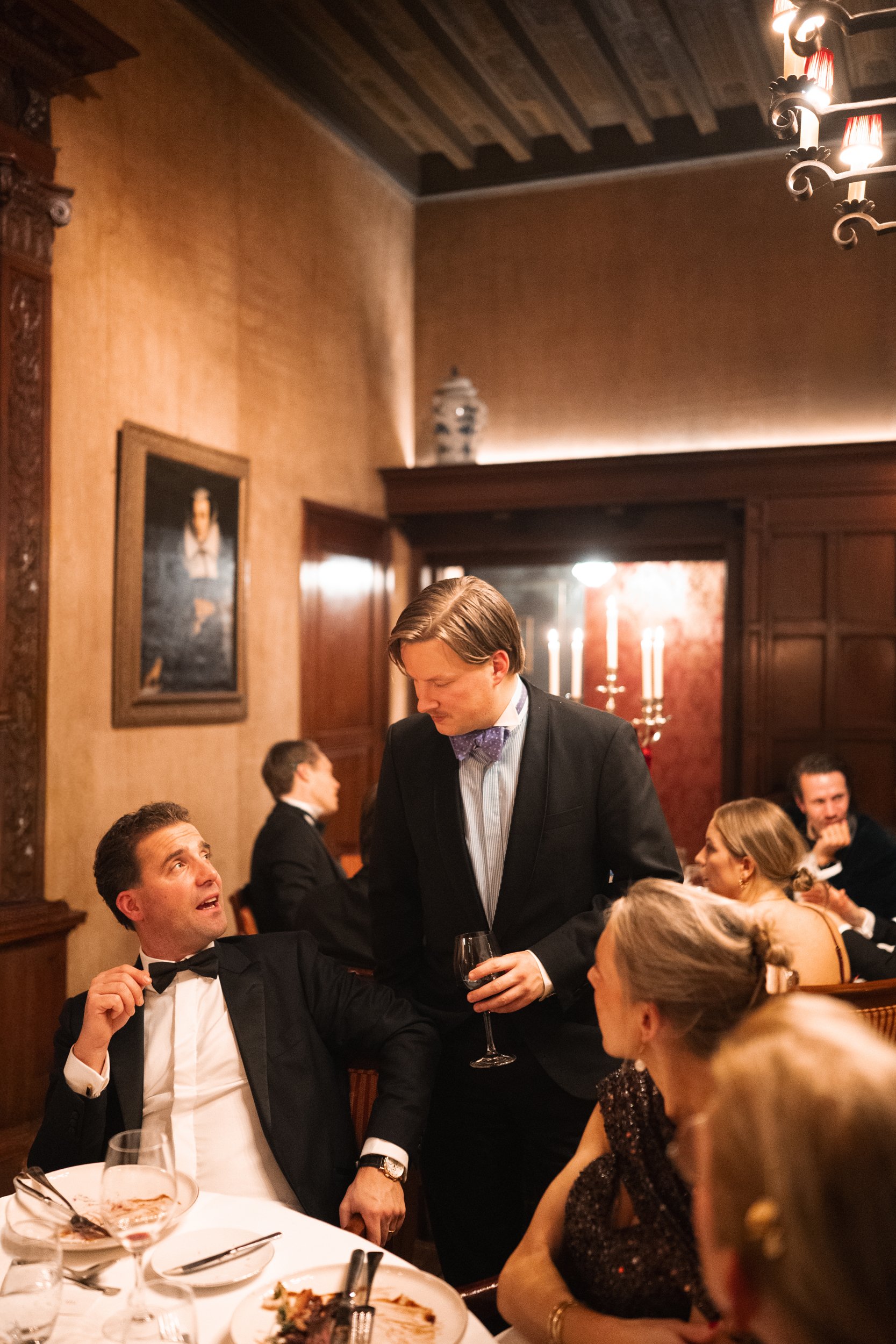 People dressed in formal attire at a dinner party in an elegant, wood-paneled room with dim lighting, a chandelier, and a portrait on the wall.