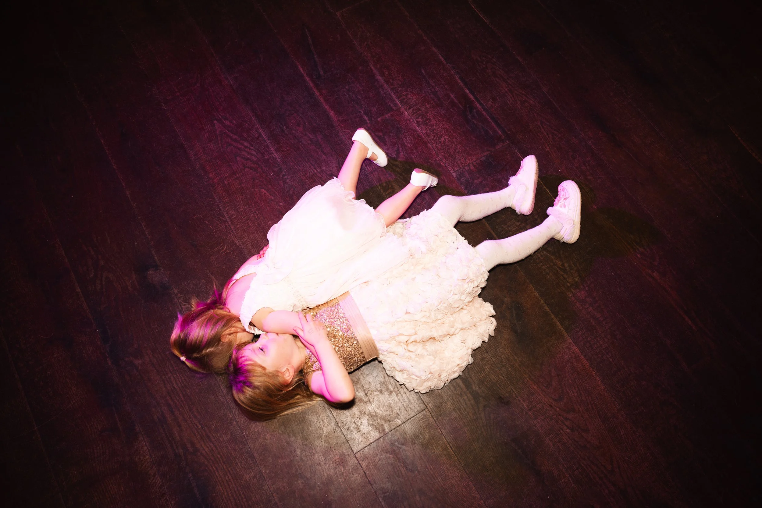 Two young girls lying on a dark wooden floor, dressed in fancy dresses, kissing each other.