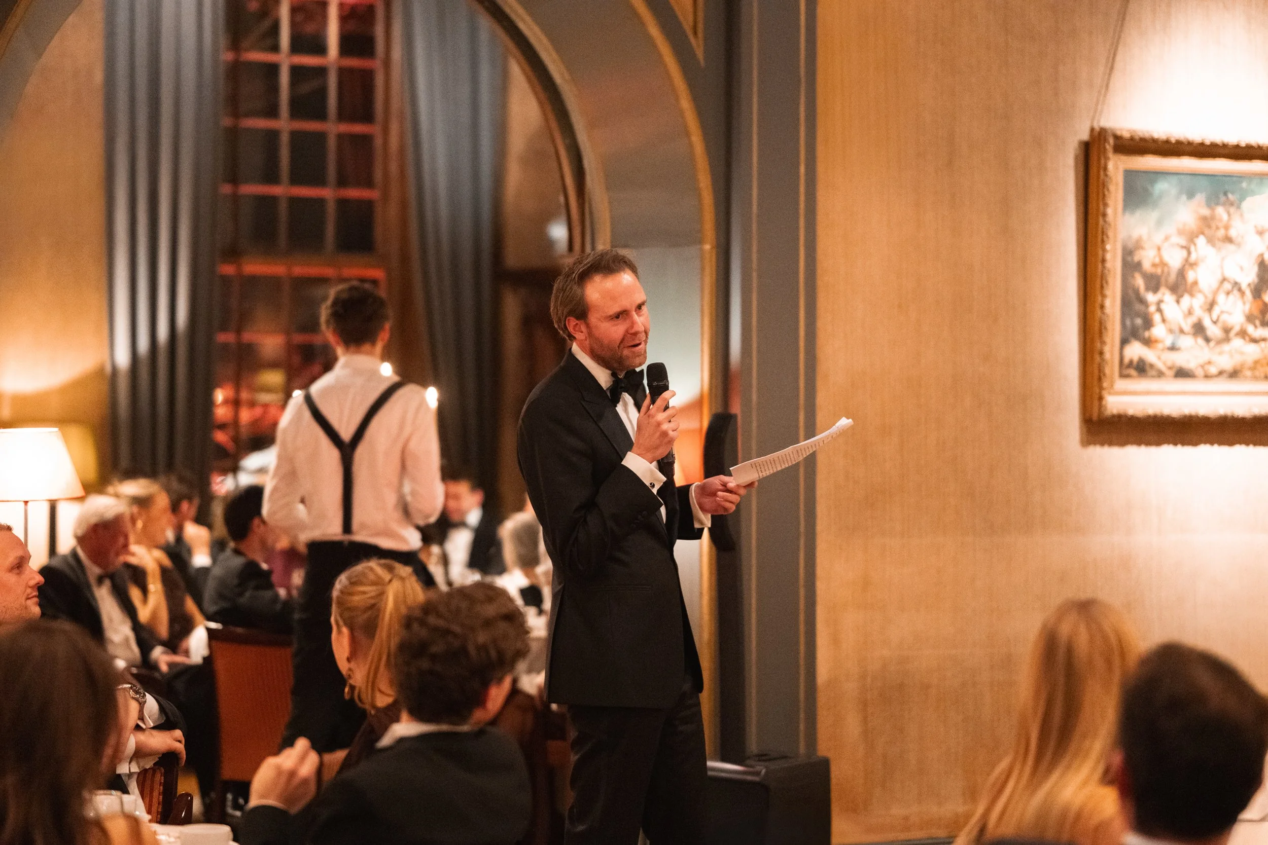 A man in a tuxedo giving a speech with a microphone at a formal dinner event in an elegant dining room.