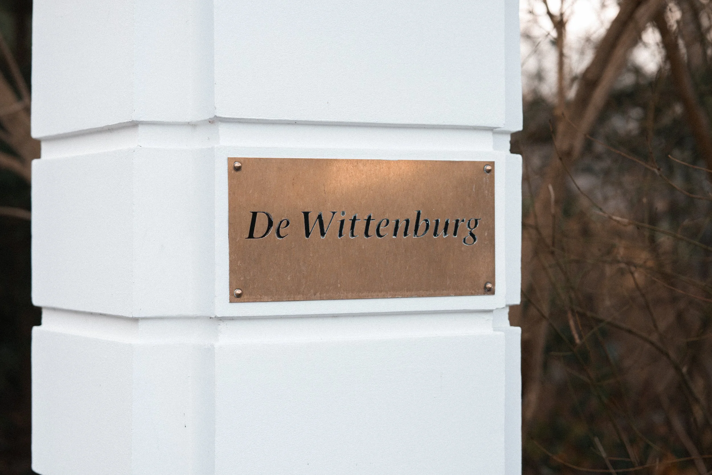 A white painted brick pillar with a brass plaque engraved with 'De Wittenburg' attached with four screws.