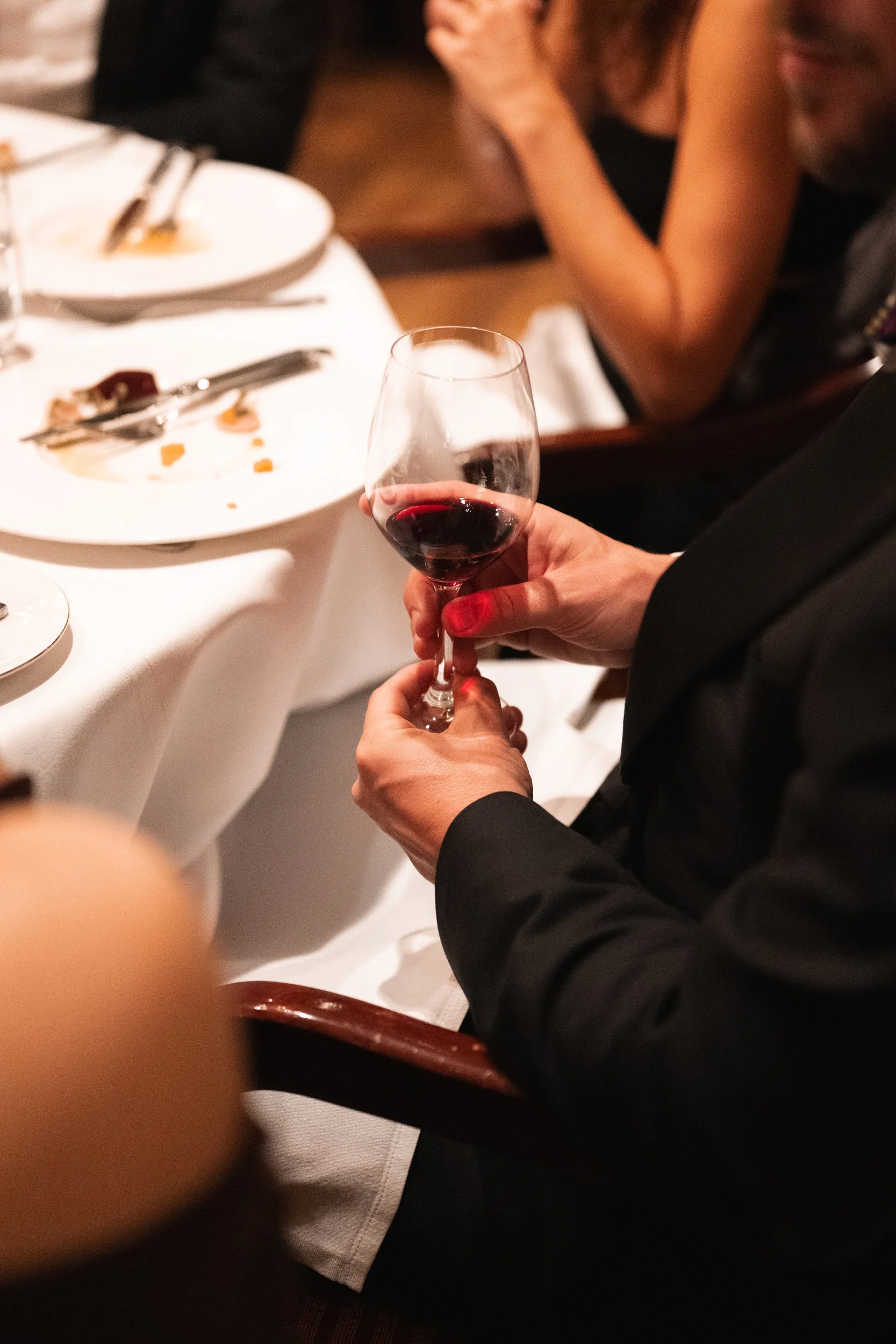 Person in a black suit holding a glass of red wine at a dinner table with dishes and utensils, with other people in the background.