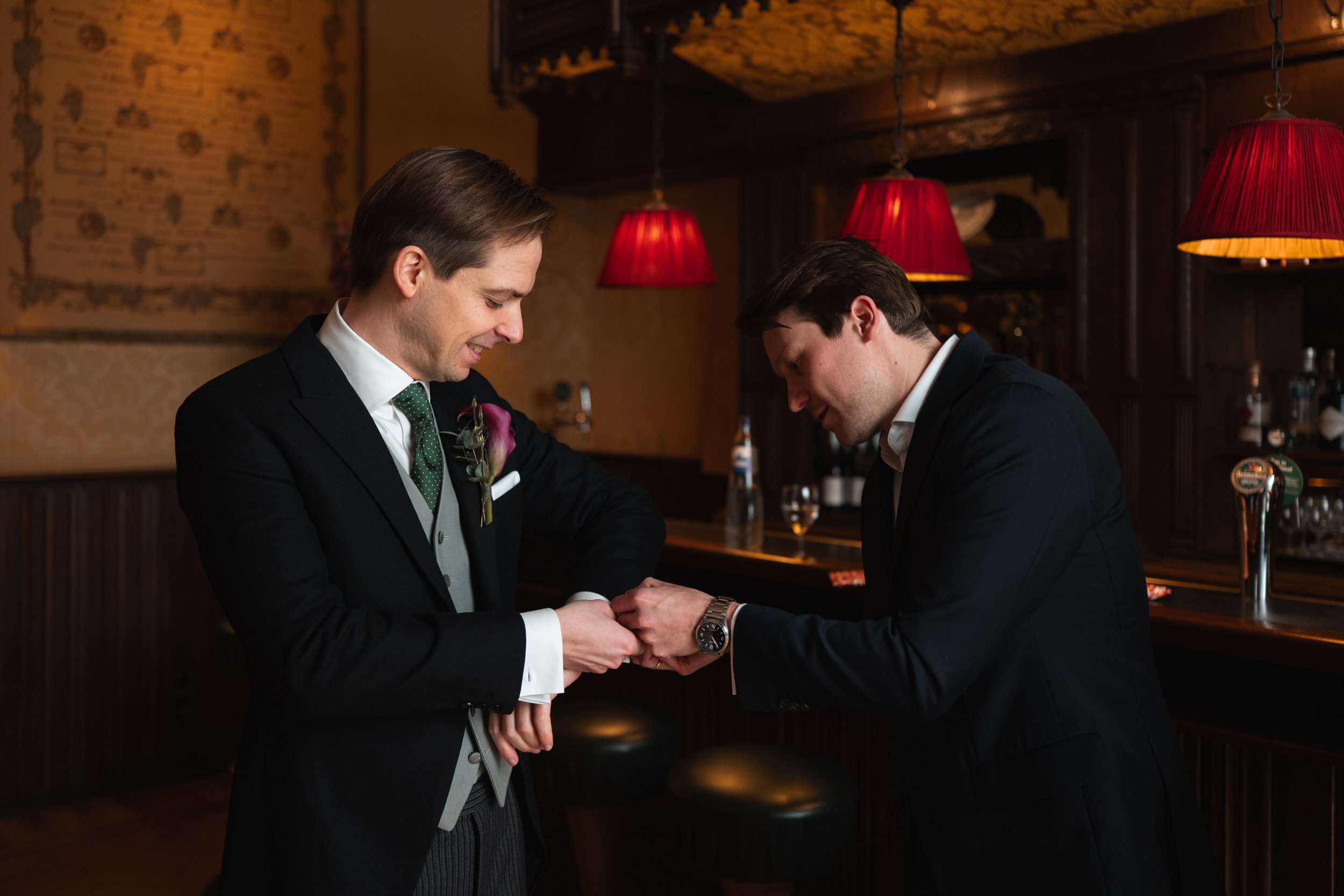 A man in a black suit hands a ring to another man in a black suit inside a dimly lit bar or pub, with red lamps hanging from the ceiling and a wooden bar counter in the background.