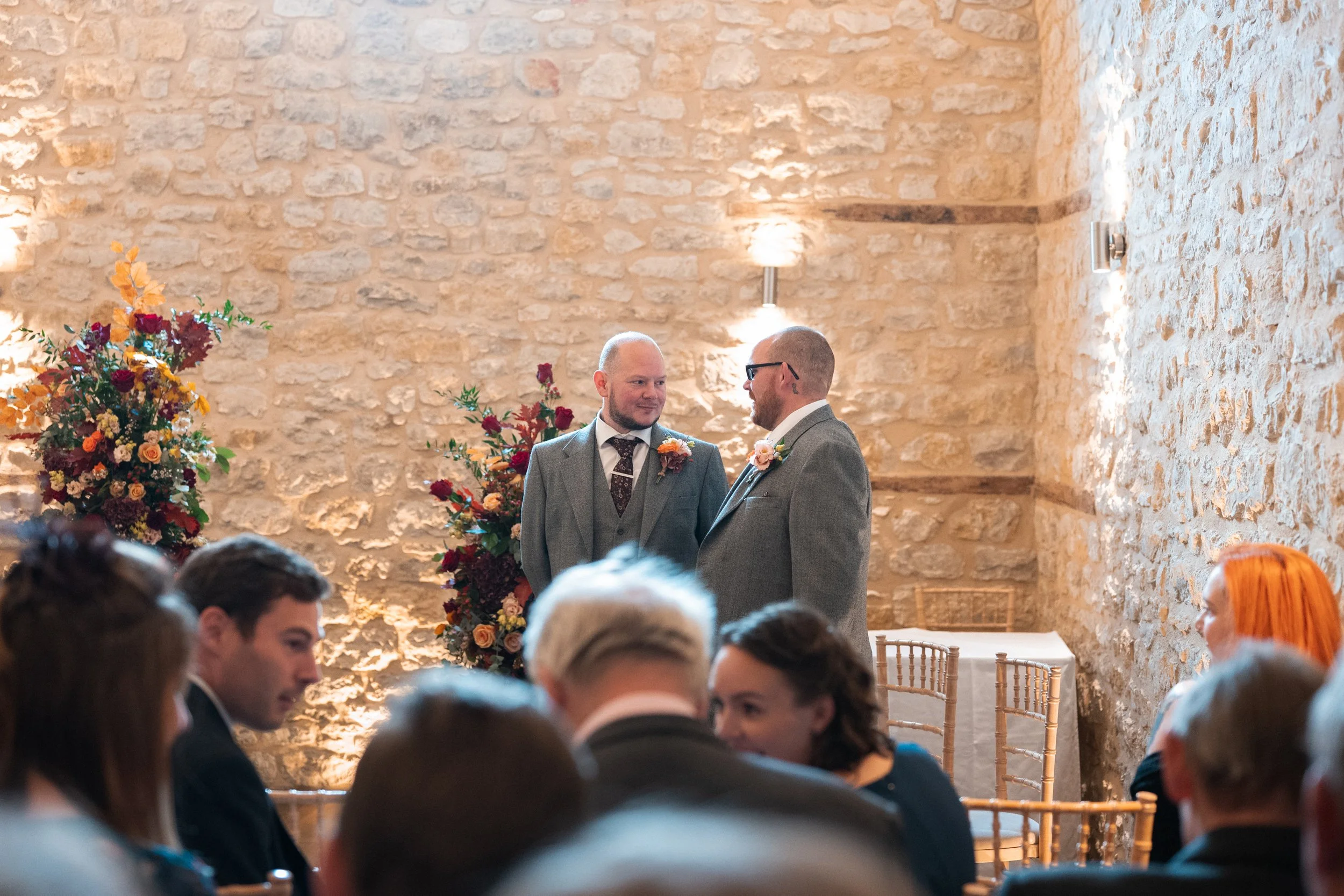 Two men in suits with boutonnières face each other and smile during a wedding ceremony inside a stone-walled venue, surrounded by seated guests and floral arrangements.