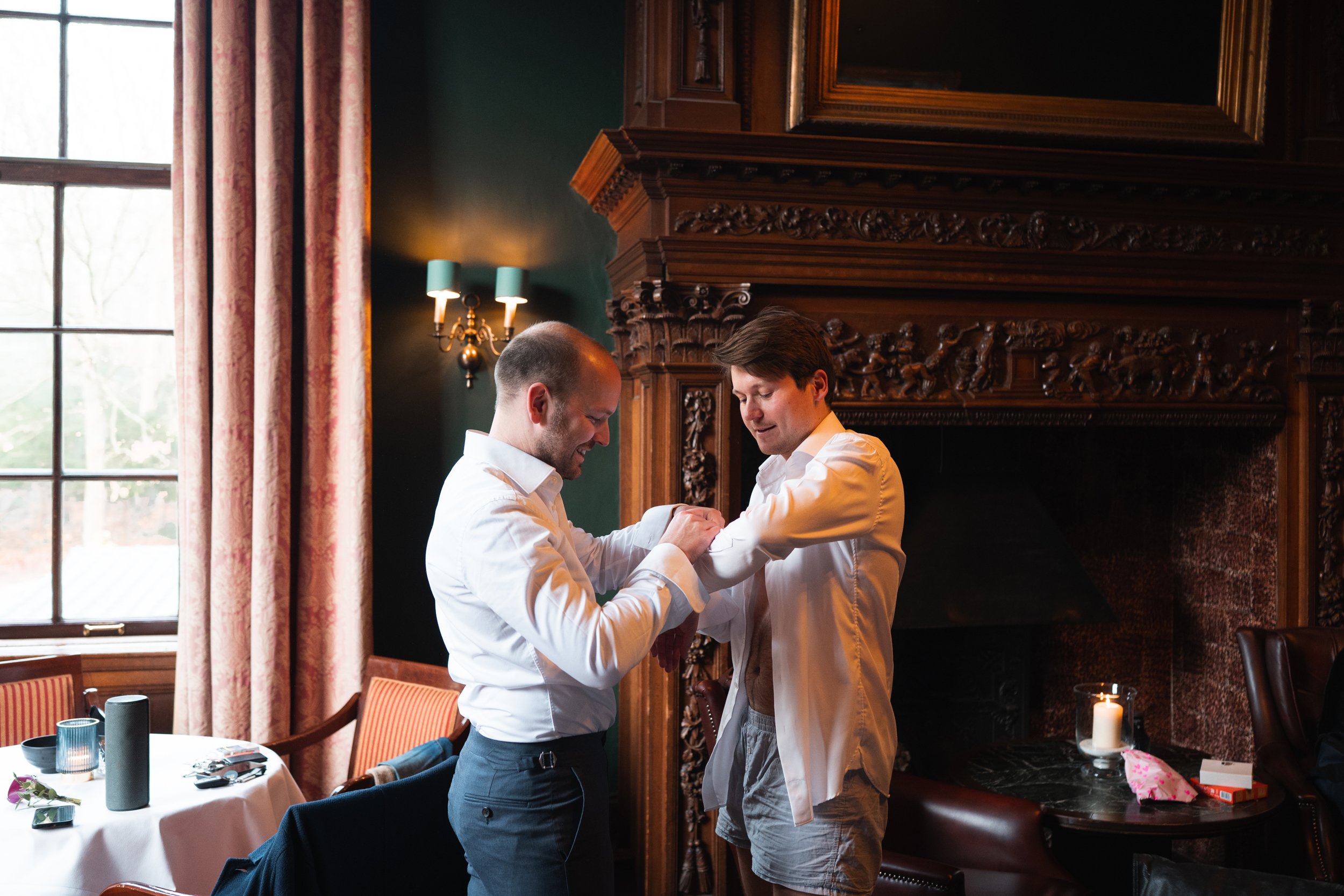 Two men in white shirts groom each other in a cozy, dimly lit room with dark green walls, ornate woodwork, a large window with curtains, and a fireplace with a candle and small gift box on nearby table.