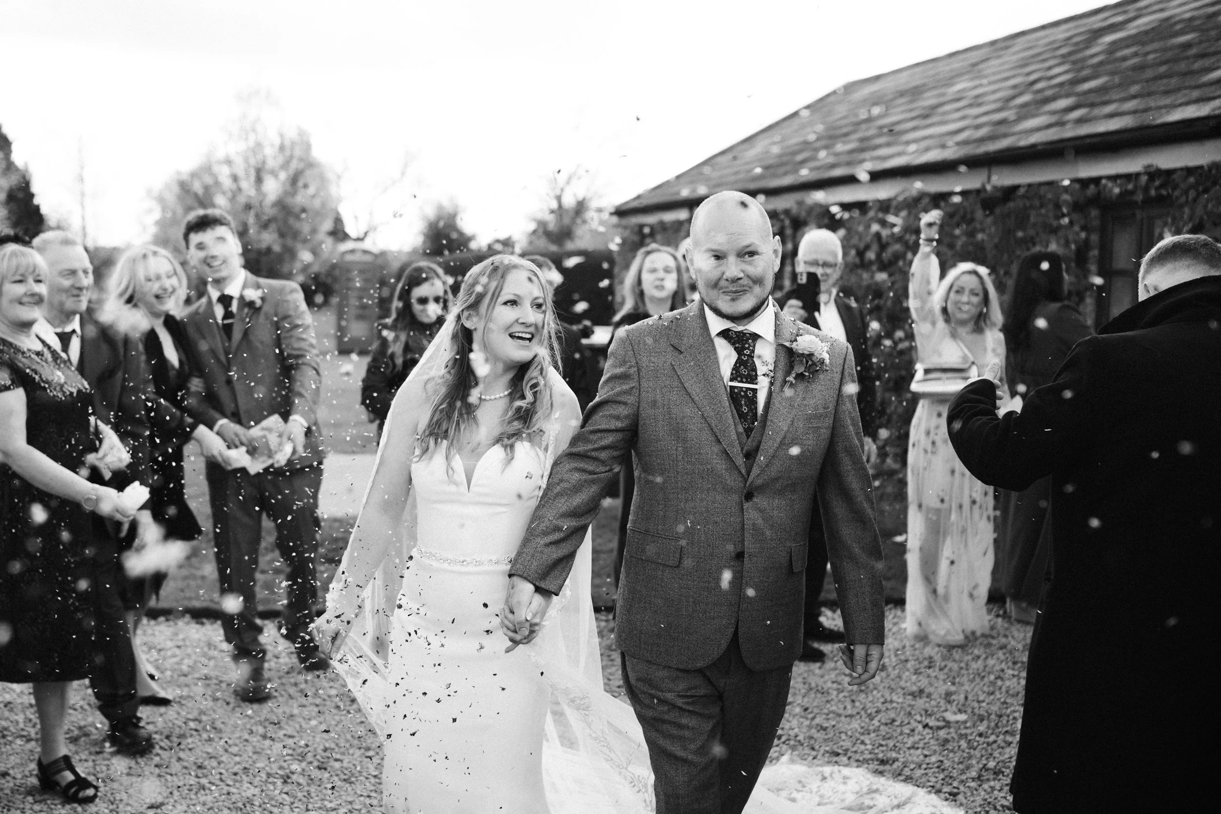 A black and white photo of a newlywed couple walking hand in hand, surrounded by wedding guests celebrating outdoors.
