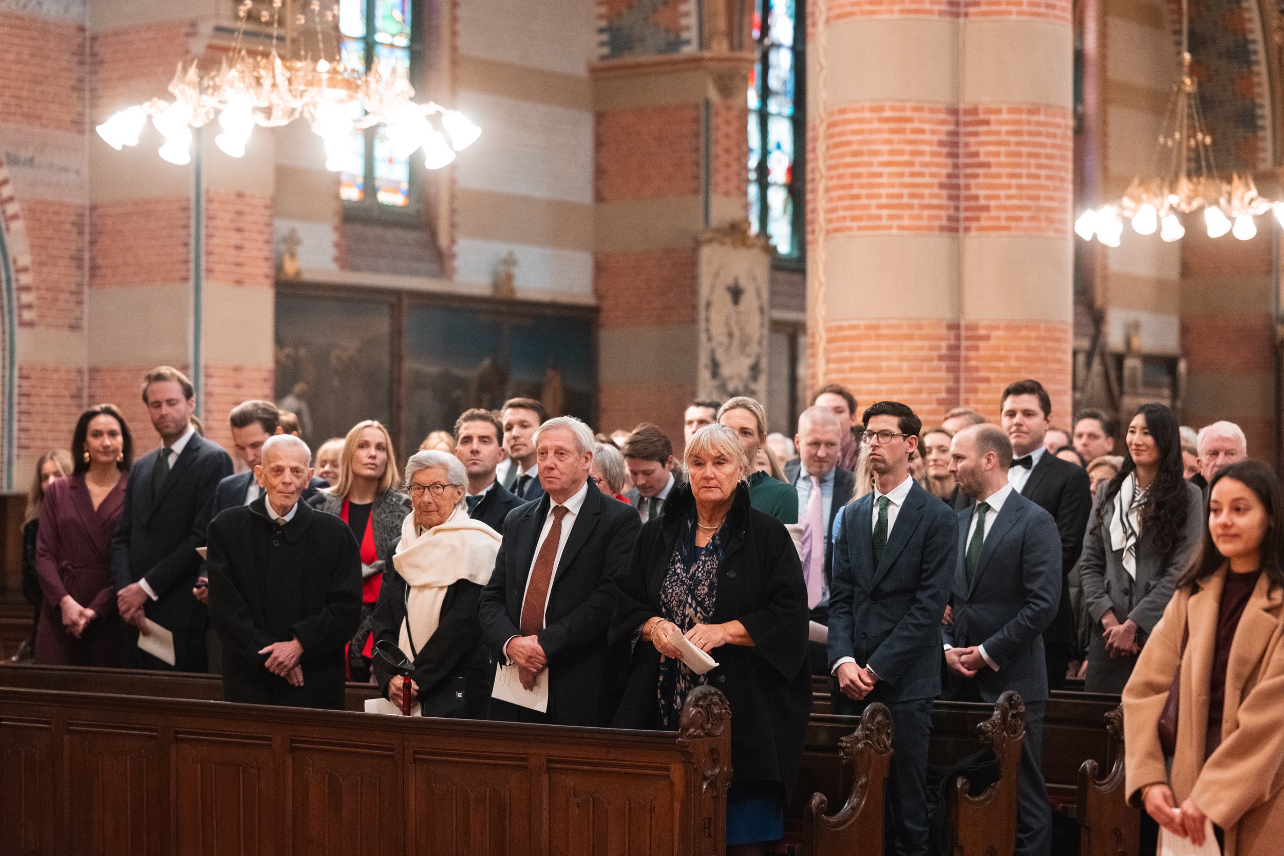 People attending a service or event in a church with stained glass windows and brick walls, standing in pews.