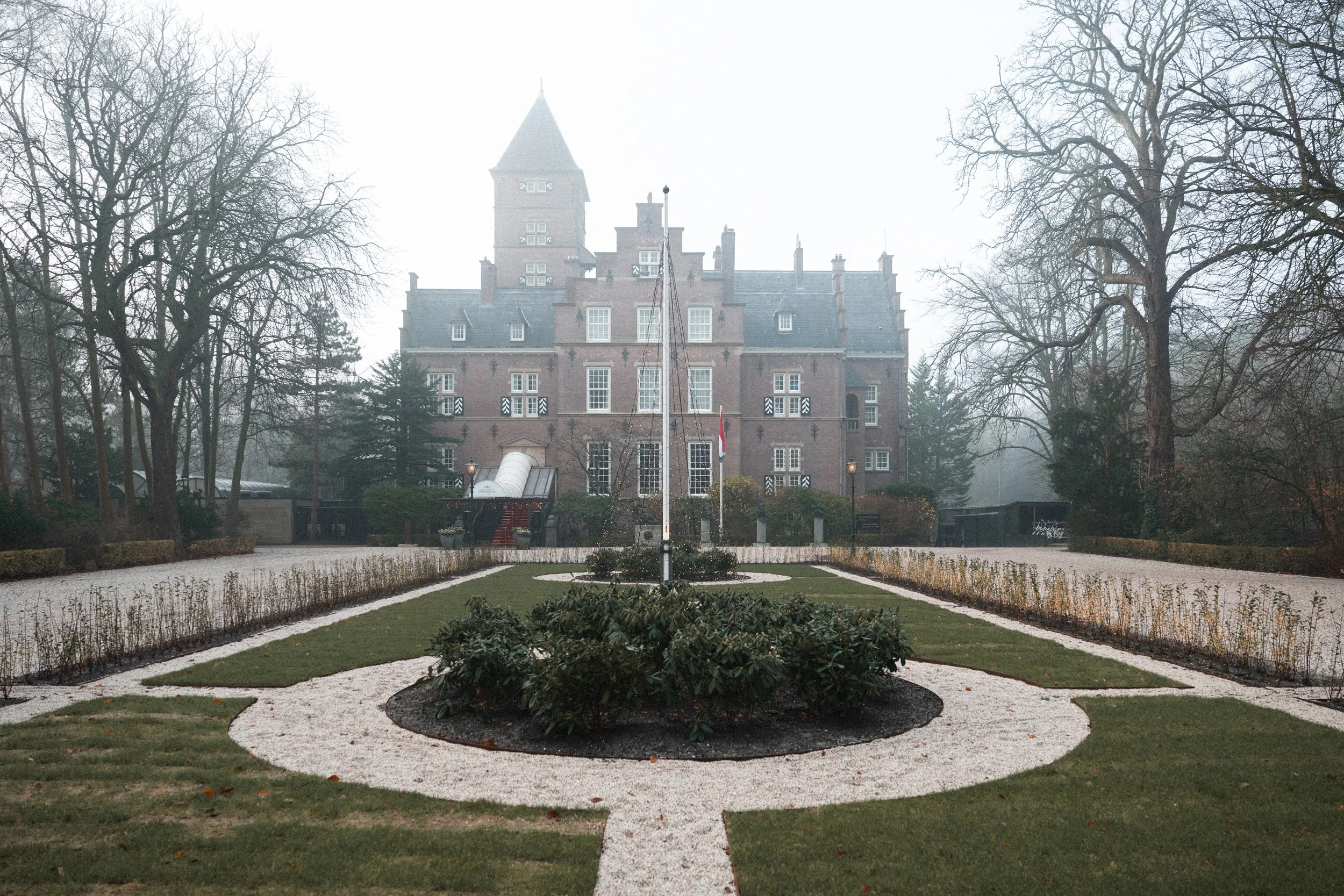 A large, historic brick castle with towers and multiple windows, surrounded by leafless trees and a well-maintained garden with paths, in foggy weather.