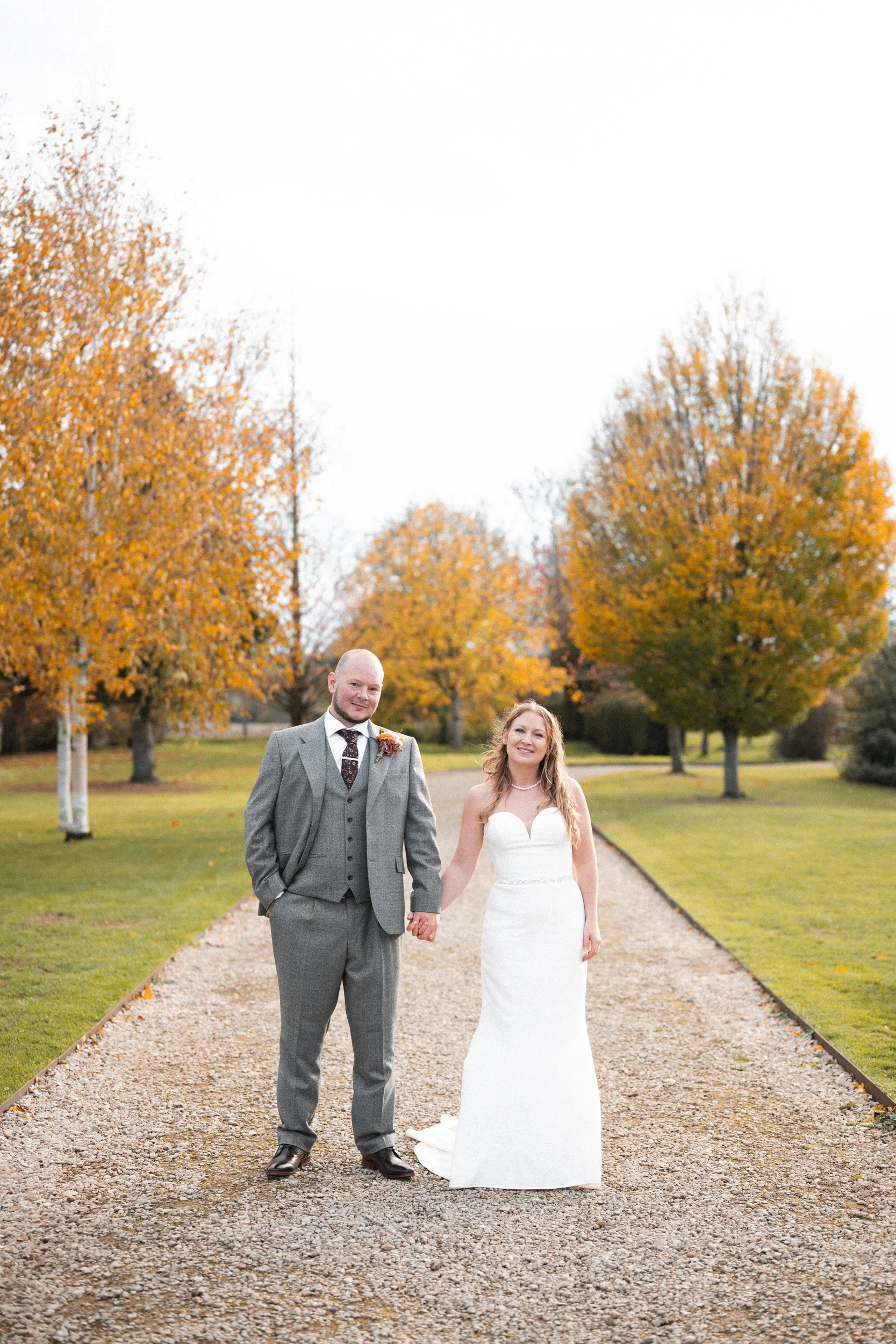 A bride and groom holding hands and smiling on a gravel path in a park with autumn trees in fall foliage.