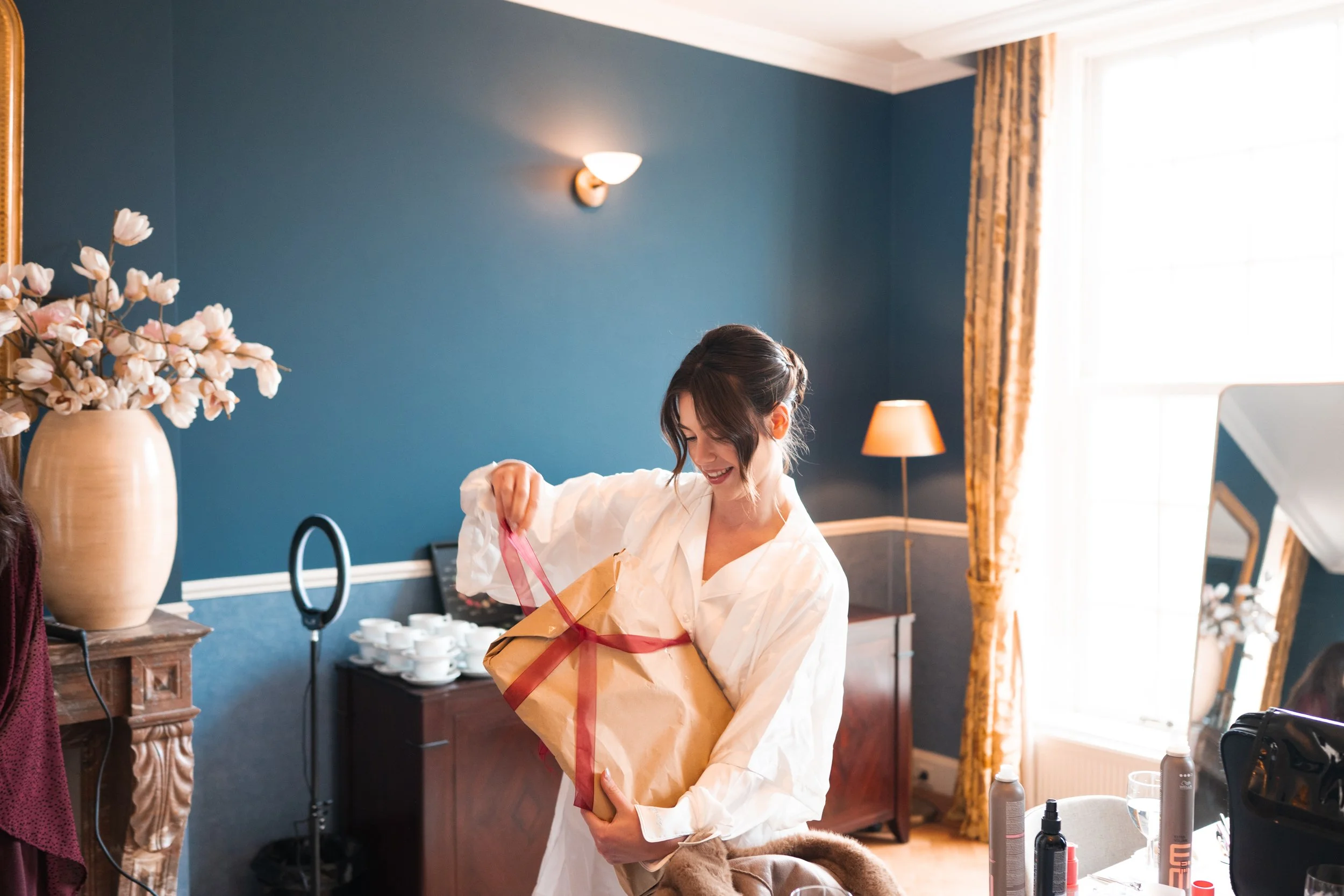 A woman in a white shirt opens a gift wrapped in brown paper with a red ribbon, smiling inside a well-lit room with blue walls, floral curtains, a large mirror, and a vase with pink flowers.