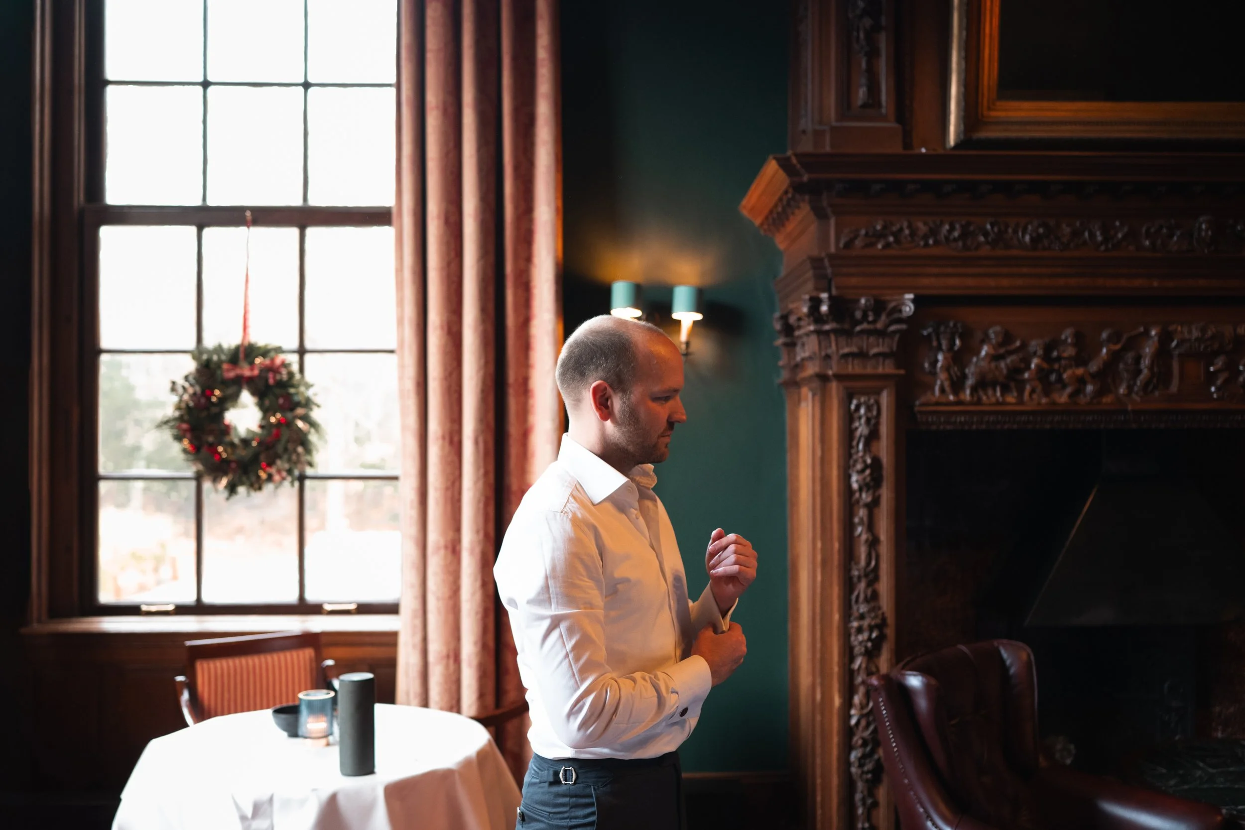 A man in a white shirt standing in a decorated room with a large window, a wreath, and ornate wooden fireplace