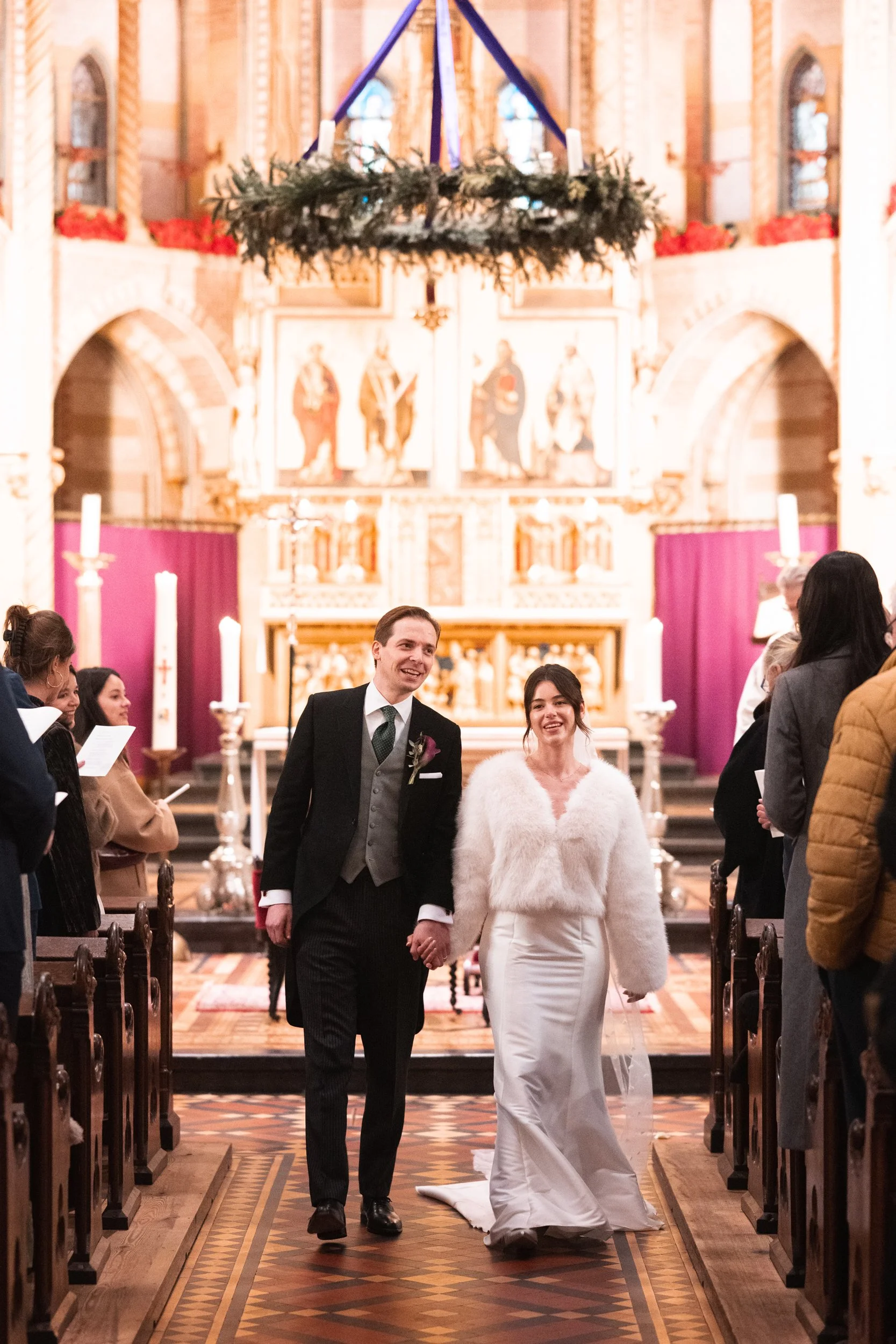 A bride and groom walking down the aisle holding hands inside a church, surrounded by guests. The bride is wearing a white wedding dress and fuzzy white sweater, while the groom is in a black suit with a gray vest and green tie. The church interior i